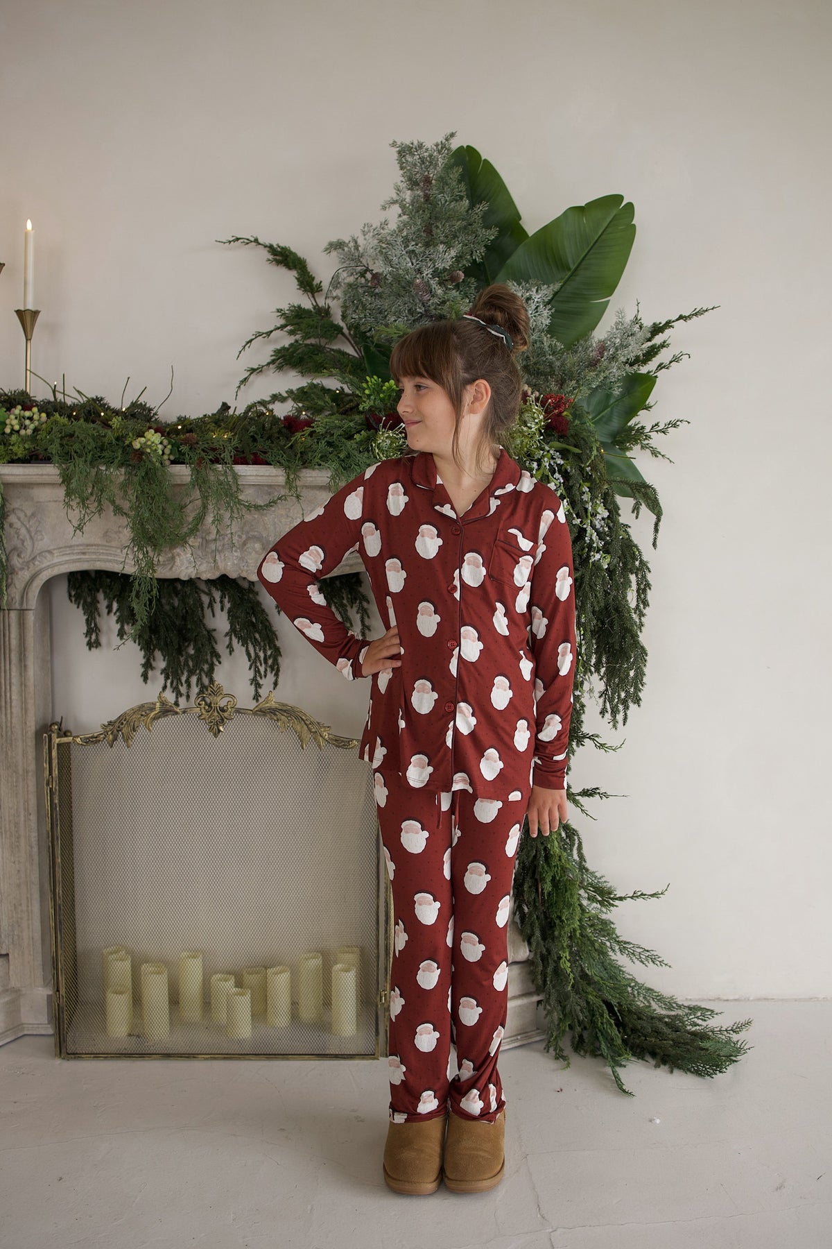 A girl wearing a dark red, long-sleeved Girl’s Bamboo Button Up Lounge Set | Vintage Santa featuring a white Santa Claus pattern on the button-up pajamas.