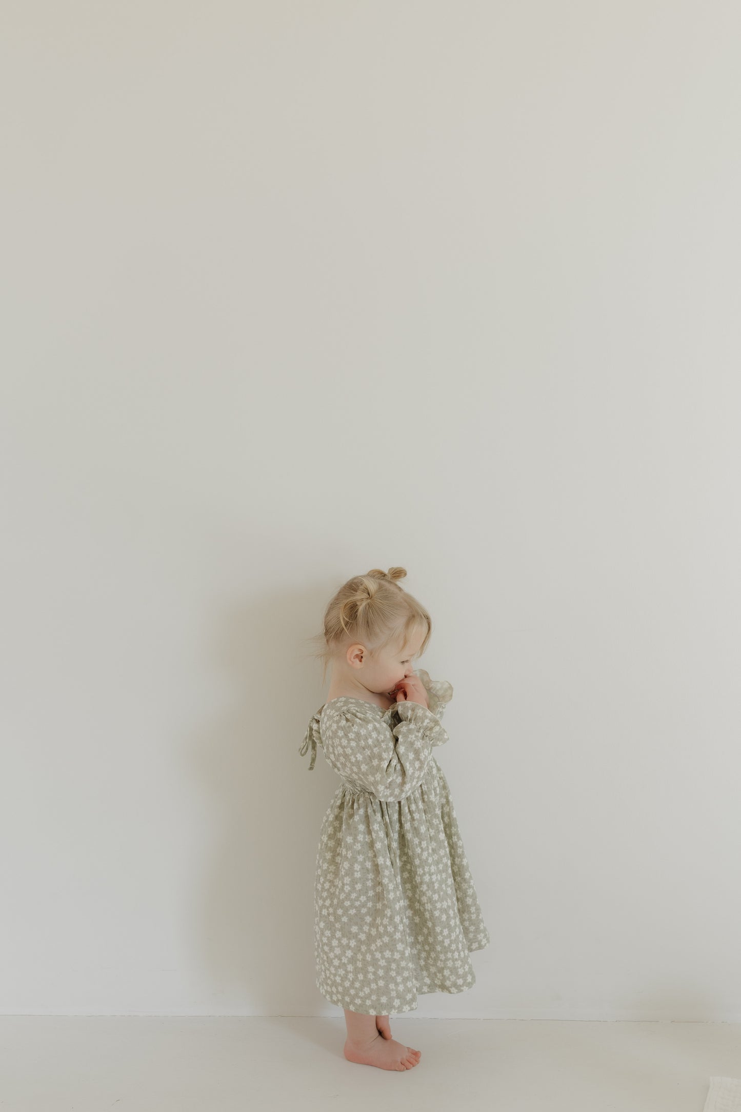 A young child with blonde hair in buns stands against a plain white wall, wearing the hypo-allergenic comfort of a "French Gray Floral" dress by forever french baby. She thoughtfully looks down, holding her hand to her mouth.