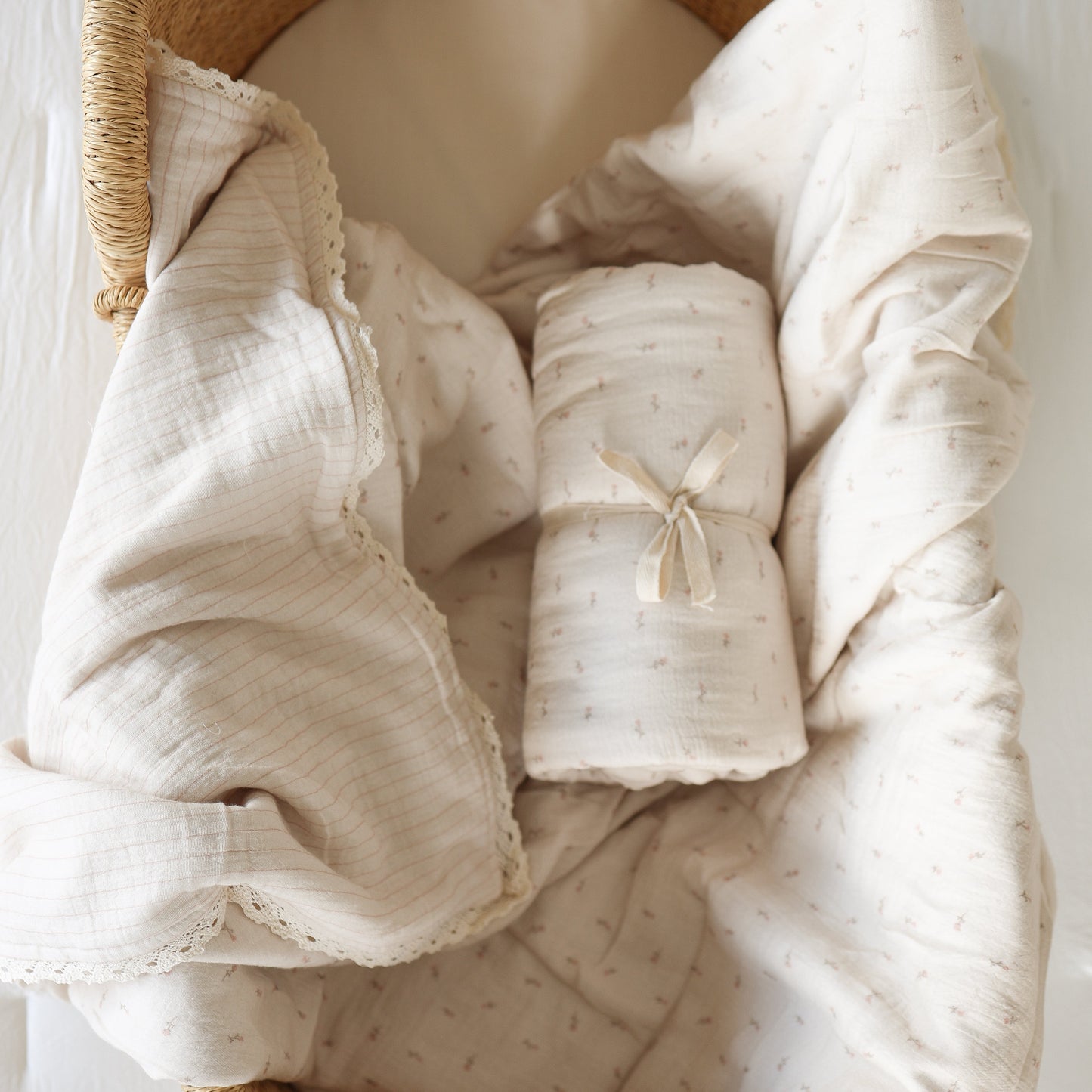 The Muslin Quilt | Lucy by forever french baby—a soft, beige 100% cotton quilt with subtle patterns—lies in a woven basket, topped with its matching rolled blanket, perfect as nursery bedding.
