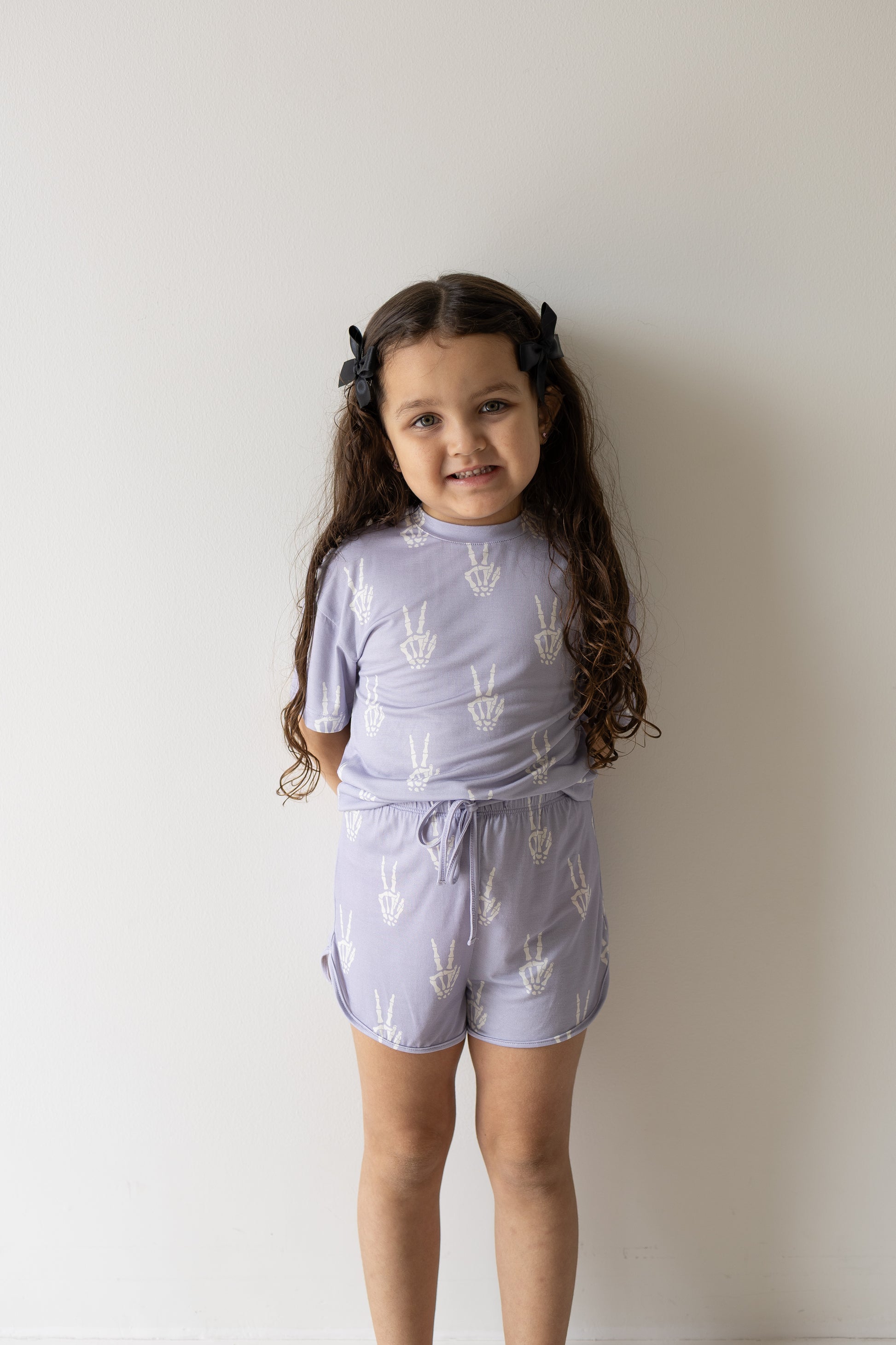 A young girl with long brown hair, dressed in the forever french baby Girl’s Bamboo Short Set | Enchanted Skelly featuring a light purple color and white peace sign patterns, stands against a plain white wall and smiles slightly. This hypoallergenic set is a stylish and comfortable choice for kids with sensitive skin.