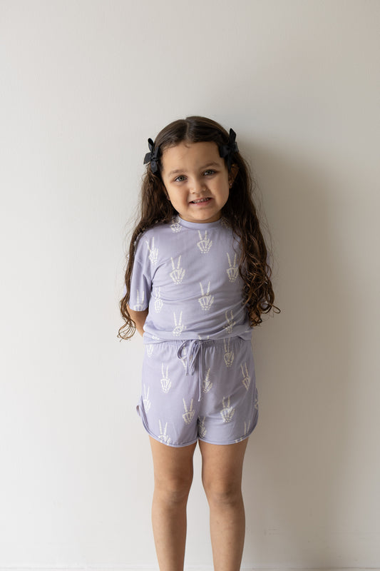A young girl with long brown hair, dressed in the forever french baby Girl’s Bamboo Short Set | Enchanted Skelly featuring a light purple color and white peace sign patterns, stands against a plain white wall and smiles slightly. This hypoallergenic set is a stylish and comfortable choice for kids with sensitive skin.