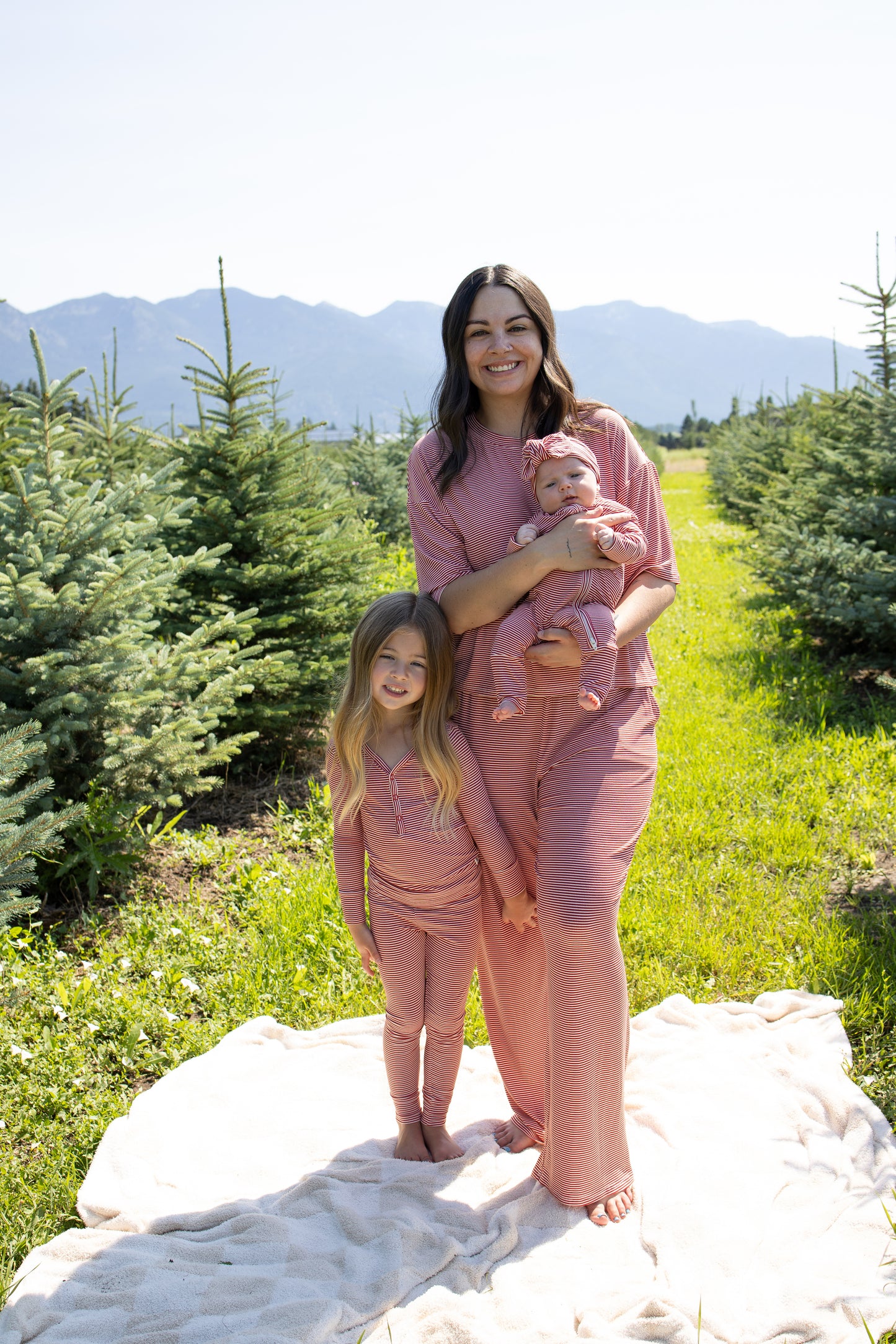 A woman stands on a blanket outdoors, holding a baby, with a young girl beside her. All three wear matching forever french baby Bamboo Two Piece Pajamas in Peppermint Stripe Ribbed, surrounded by small pine trees and distant mountains under a clear sky.