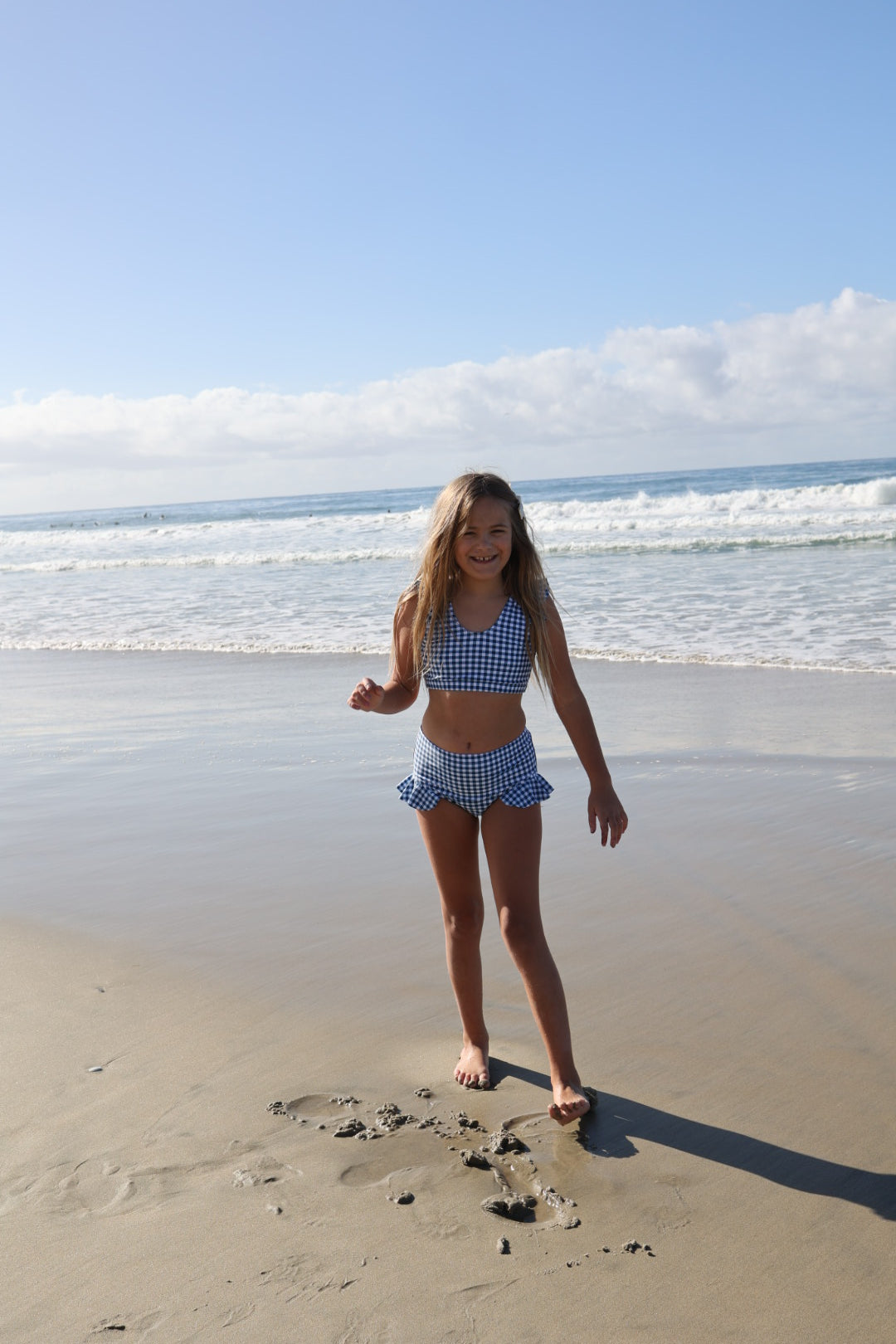 A young girl wears the forever french baby Girls Two Piece Swimsuit in Blue Gingham, smiling on a sandy beach with gentle waves and a blue sky behind her.