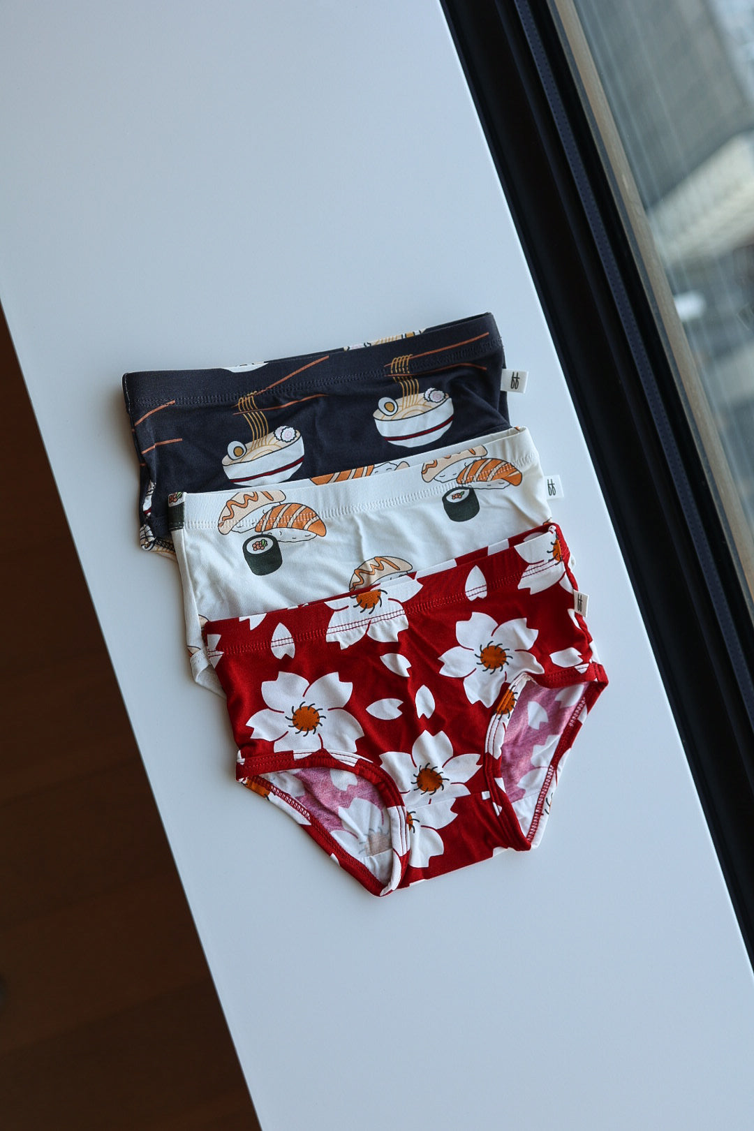 A set of three forever french baby Girl's Bamboo Briefs (Cherry Blossoms, Ramen & Sushi) is neatly stacked on a white surface by a window, featuring playful ramen and sushi prints plus bold cherry blossoms.