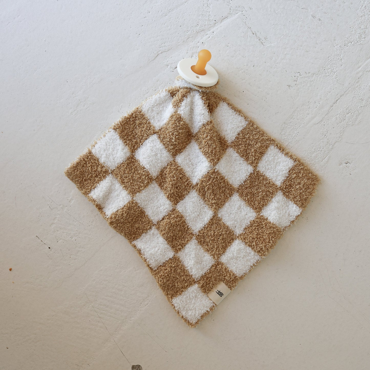 The Lovey | Original Checkerboard from forever french baby, a plush microfiber baby blanket with a beige and white checkered pattern and attached pacifier, rests on a textured white surface.
