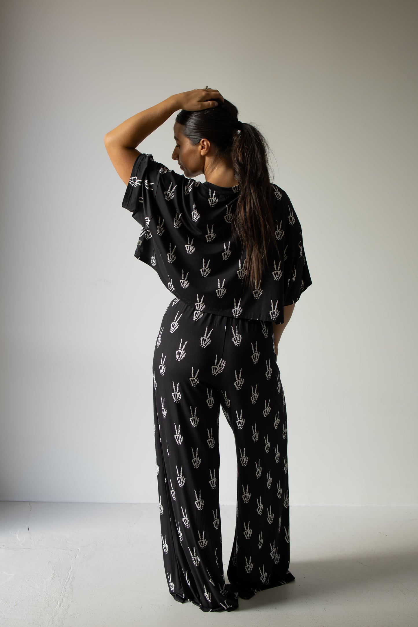 A person with long dark hair in a ponytail stands facing away, one arm raised to touch their head, wearing the forever french baby Women’s Bamboo Crop Tee Lounge Set in Midnight Skelly—a loose black bamboo viscose crop tee featuring a white handprint pattern paired with wide-leg pants. The soft and breathable bamboo fabric enhances the relaxed, effortless vibe in the minimalist room with pale walls and floor.