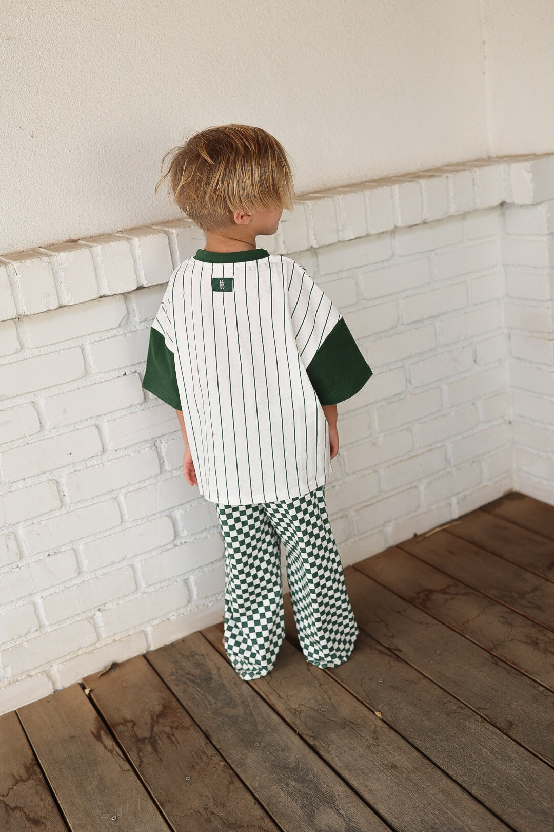 A blonde-haired child stands on a wooden floor facing a white brick wall, wearing the forever french baby T-Shirt & Pant Set | Lucky Micro Check with a white and green striped shirt and green-and-white checkered wide leg pants.