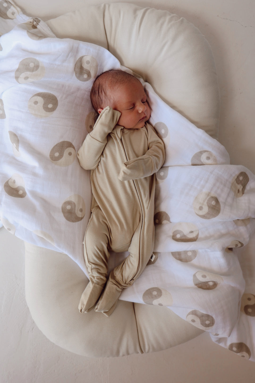 A newborn baby dressed in "Bamboo Zip Pajamas | Limestone" from forever french baby sleeps on a cushioned surface with a white and beige blanket featuring yin-yang symbols partially covering them. The baby has one hand touching their face and is surrounded by a calm, cozy setting.