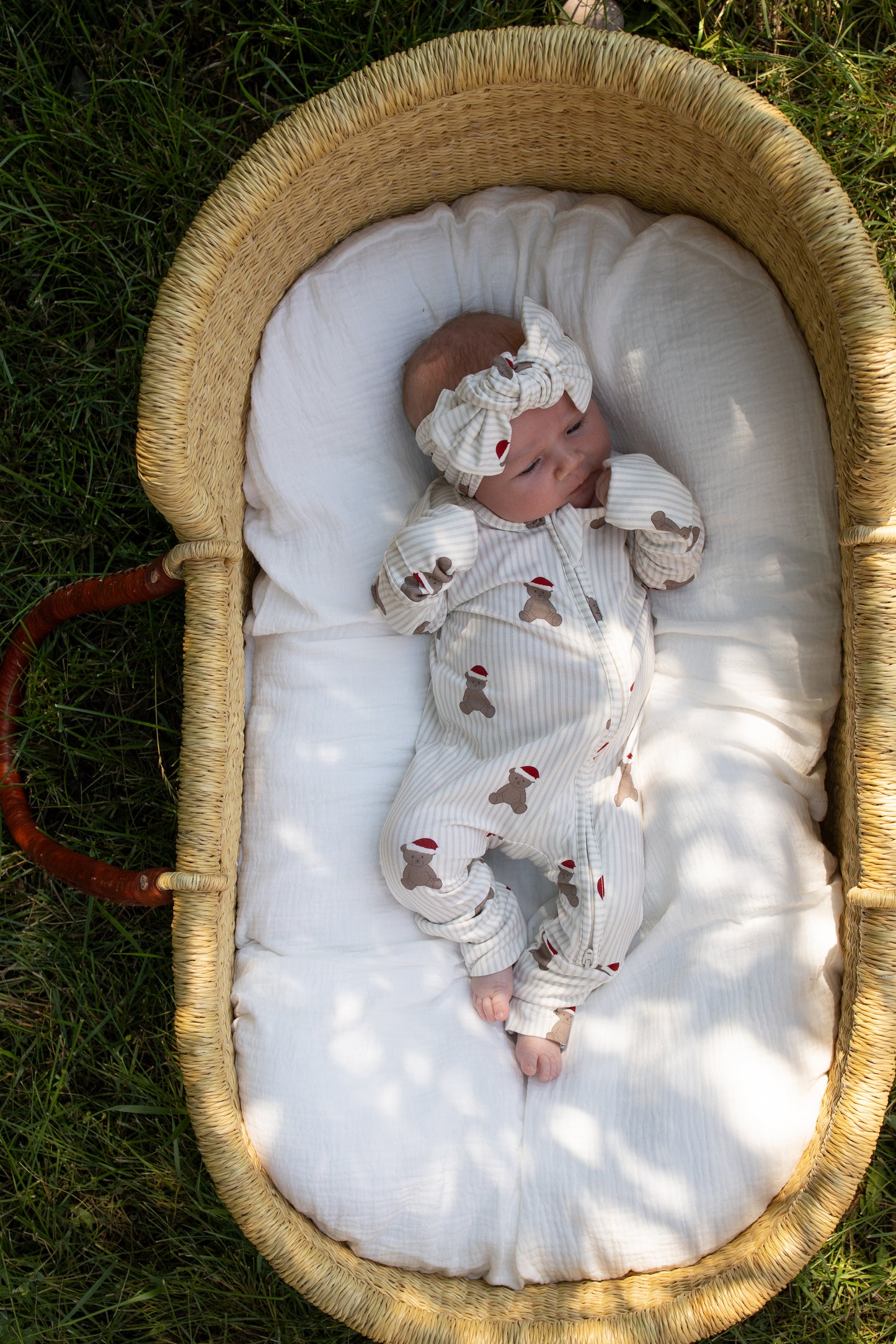 A baby in the Bamboo Zip Pajamas | Santa Teddy from forever french baby, complete with a matching headband, lies in a woven basket on a white cushion, surrounded by grass and dappled sunlight. Crafted from hypoallergenic materials, this cozy outfit ensures comfort and gentle care for delicate skin.