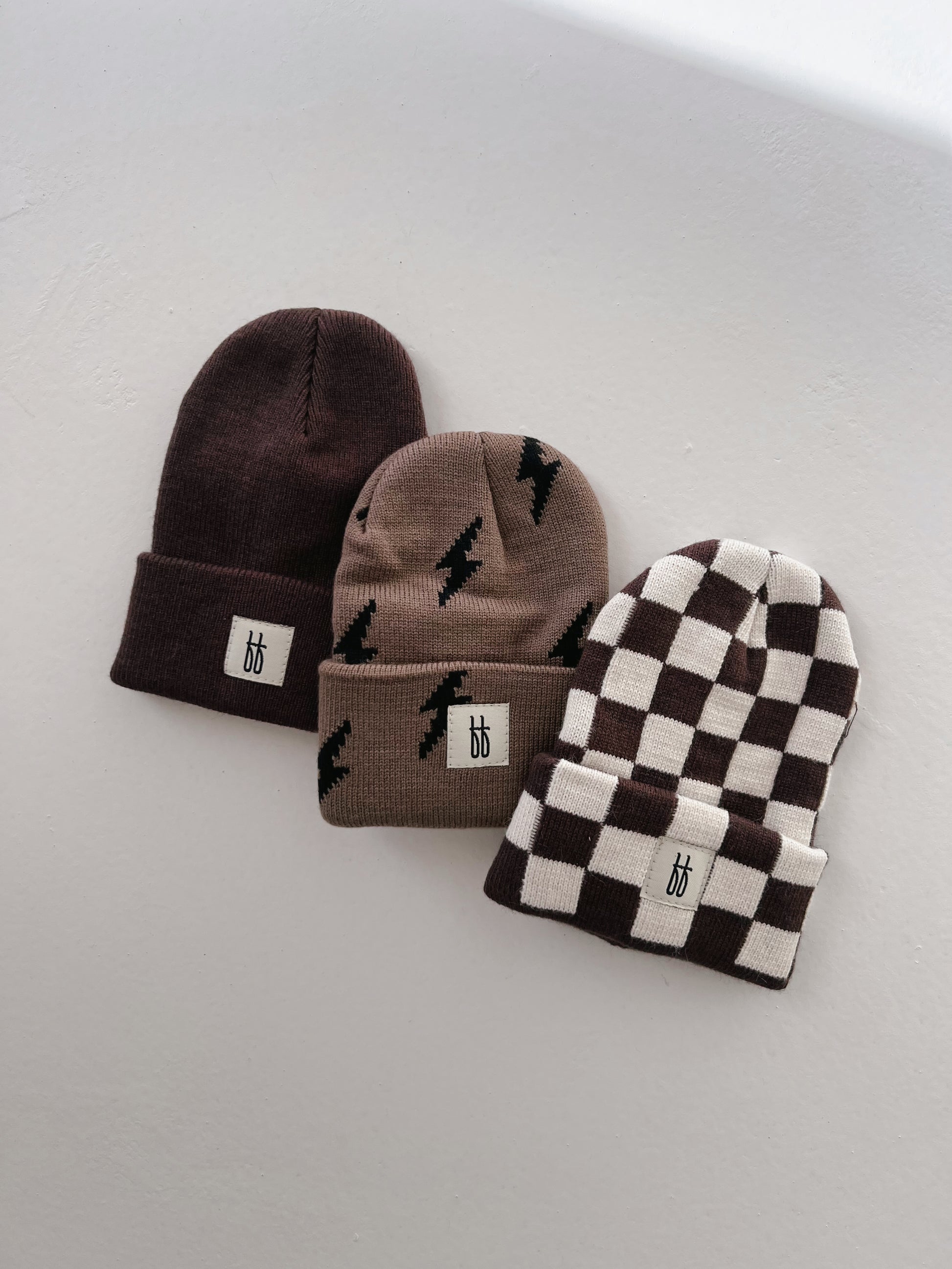 Three knit beanies on a white surface showcase different styles. The left ff Knit Beanie from forever french baby features a dark brown ribbed design, the middle beanie is beige with black lightning bolt patterns, and the right beanie sports a checkered pattern with brown and white squares. All beanies have a small white label.