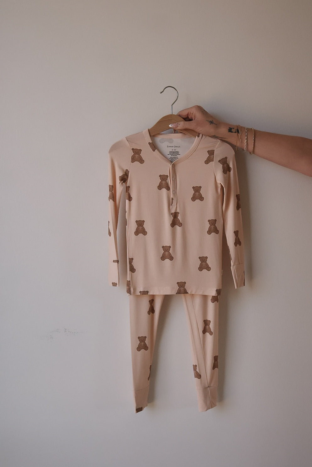 A hand displays the forever french baby Bamboo Two Piece Pajamas | Lottie—beige, hypoallergenic kids’ pajamas with a brown teddy bear print, featuring a long-sleeve buttoned top and matching pants on a hanger against a plain wall.