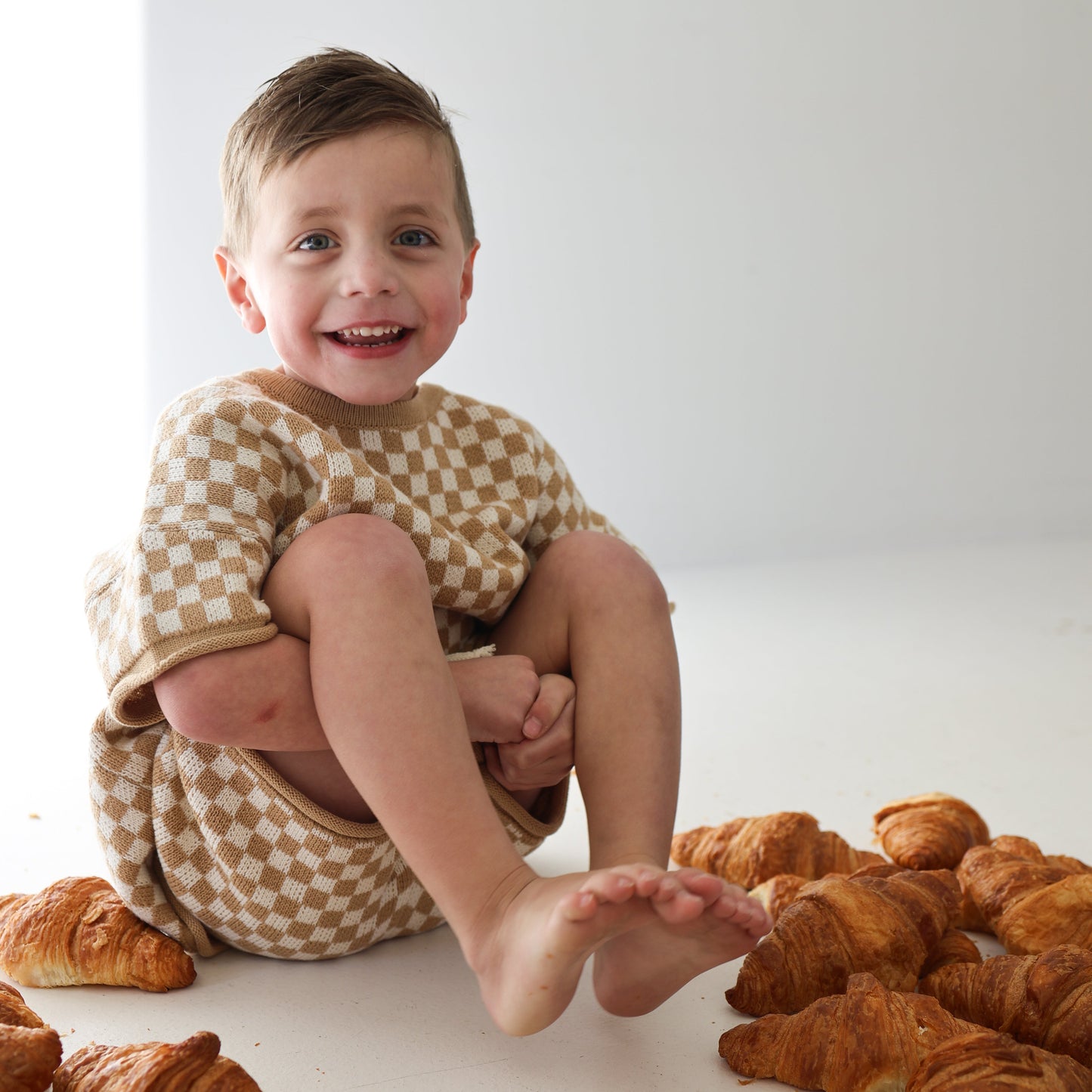 A young child with short brown hair, barefoot and smiling, wears the Knit Short Set in Latte Check from forever french baby while sitting among croissants on a white floor against a plain light background.