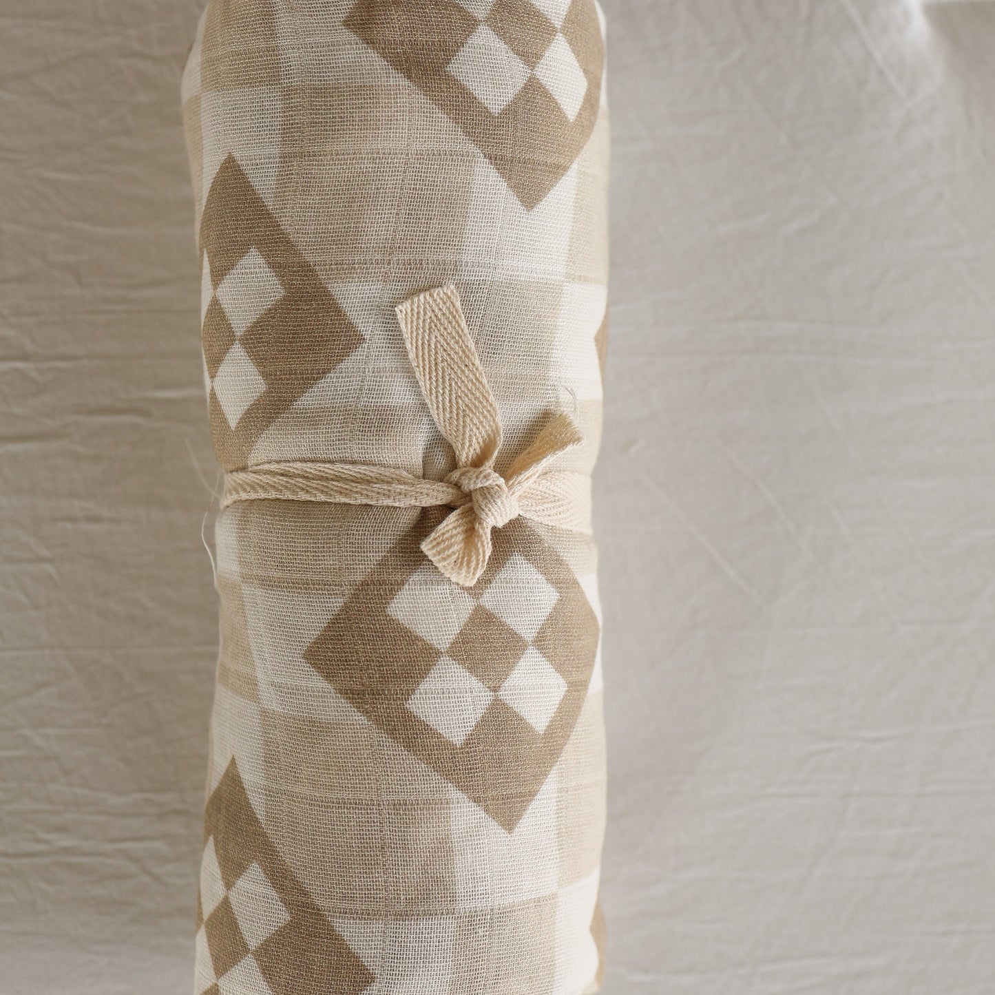 The forever french baby Muslin Swaddle | Cottage Quilt features a beige and white geometric diamond pattern, secured with a knotted beige tie, and is displayed on a textured light background.
