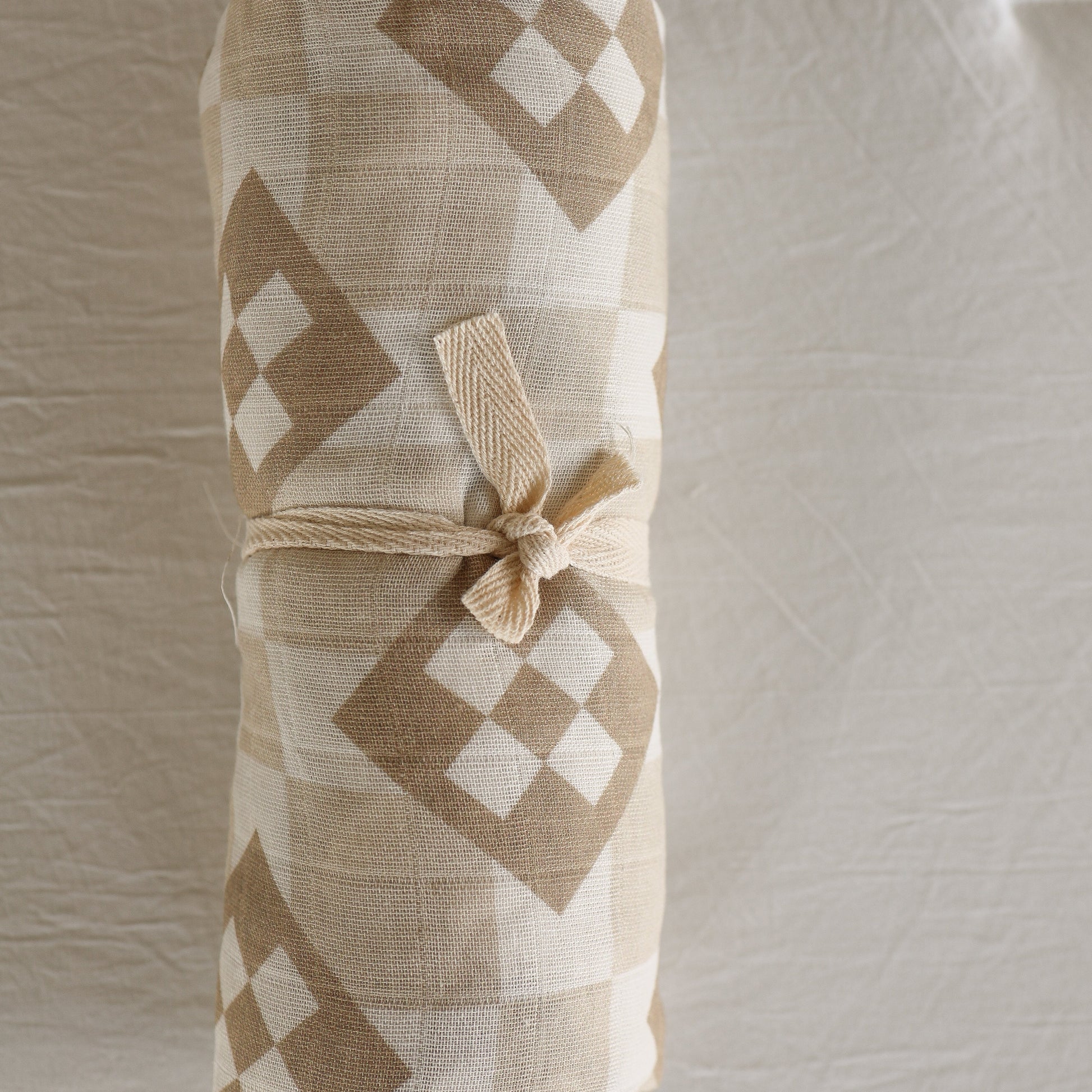The forever french baby Muslin Swaddle | Cottage Quilt features a beige and white geometric diamond pattern, secured with a knotted beige tie, and is displayed on a textured light background.