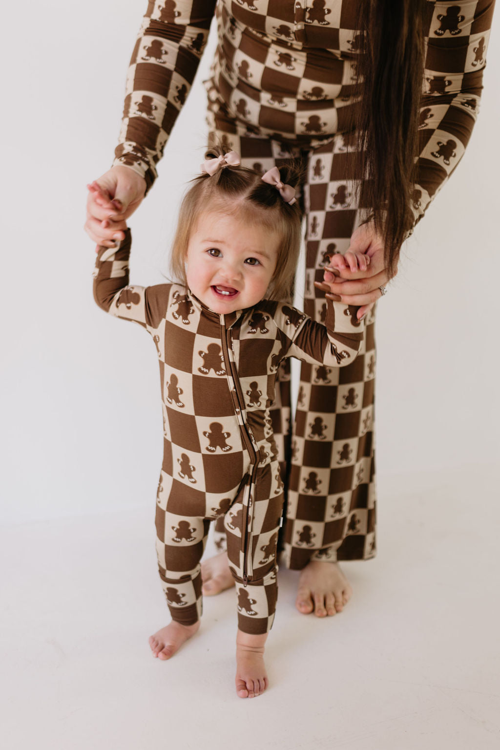 A toddler dressed in the Gingerbread Checkerboard Bamboo Zip Pajamas by forever french baby holds hands with an adult wearing matching breathable babywear. The child's hair is adorned with bows, and both are barefoot on a plain, light-colored background.