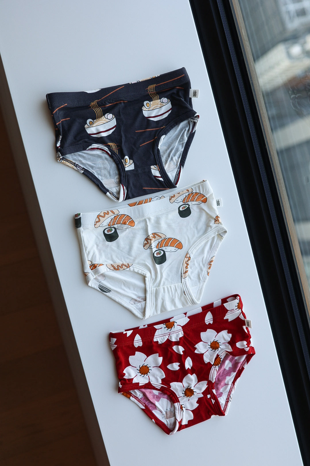 Three pairs of forever french baby Girl's Bamboo Briefs are displayed by a window: one with cherry blossoms, one with ramen, and one with sushi prints.