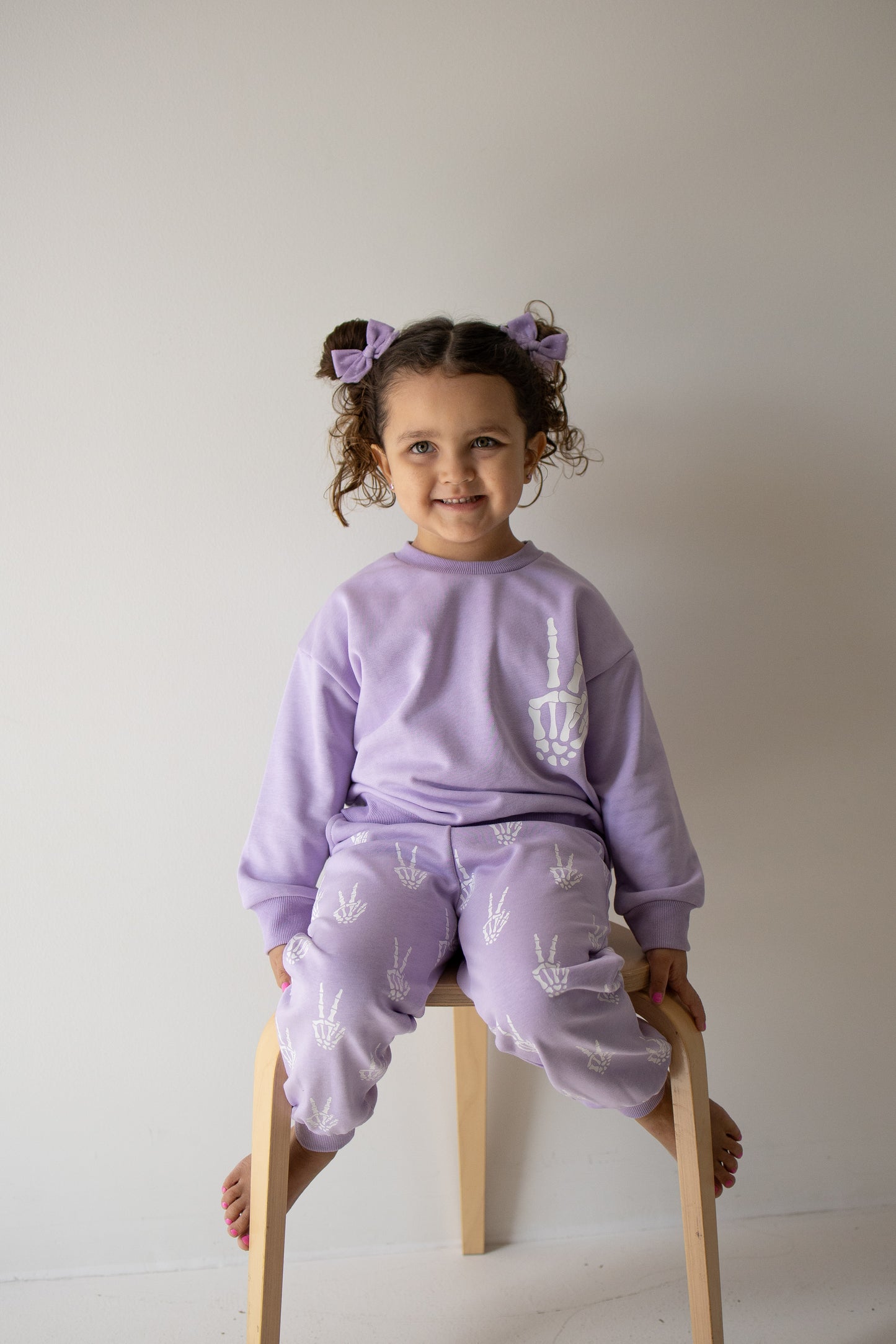 A young child with curly hair and purple bows sits on a stool, smiling and wearing the forever french baby Child Sweat Set | Enchanted Skelly—a matching lavender children’s sweat set with white designs—perfect for everyday adventures, against a plain light-colored wall.