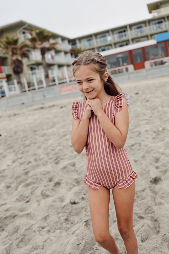 A young girl, wearing a pink and white UPF 50+ striped One Piece Girls Swim Suit from forever french baby, stands on a sandy beach with her hands clasped near her chin. She has a content expression on her face. Buildings with balconies and palm trees are visible in the background.