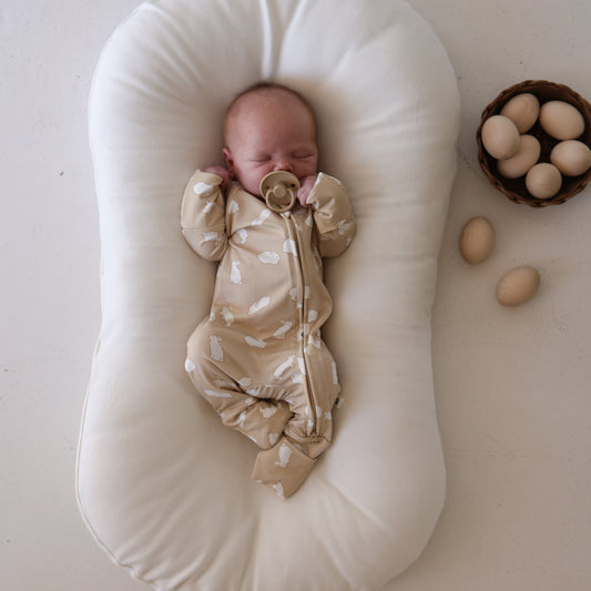 A baby in forever french baby Bamboo Zip Pajamas | Bunnies lies on a white lounger, holding a pacifier, with a basket of eggs and loose eggs nearby on a light background.
