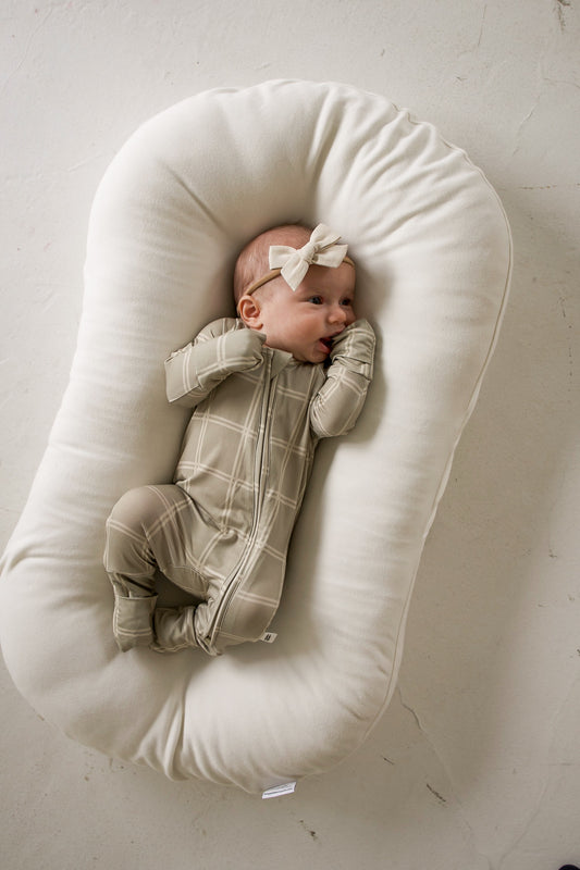 A baby wears Bamboo Zip Pajamas | Linen Grid with a beige grid-patterned zipper while lying in a cushioned lounger.