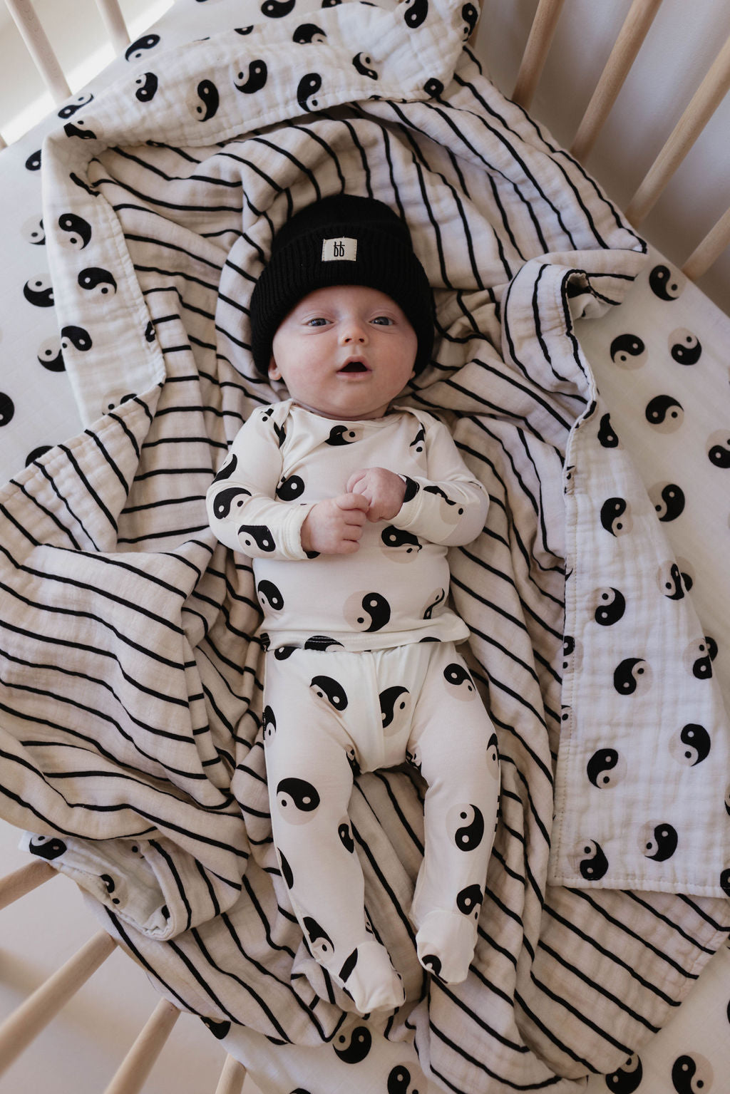 A baby lies in a crib, wearing a hypo-allergenic white onesie and cap featuring a black-and-white yin-yang pattern from the forever french baby collection. The baby is also wrapped in the matching Sleepy Time Set | Black & White Yin Yang, complemented by a striped blanket made of bamboo fabric. The wooden slats of the crib frame the baby's relaxed upward gaze.