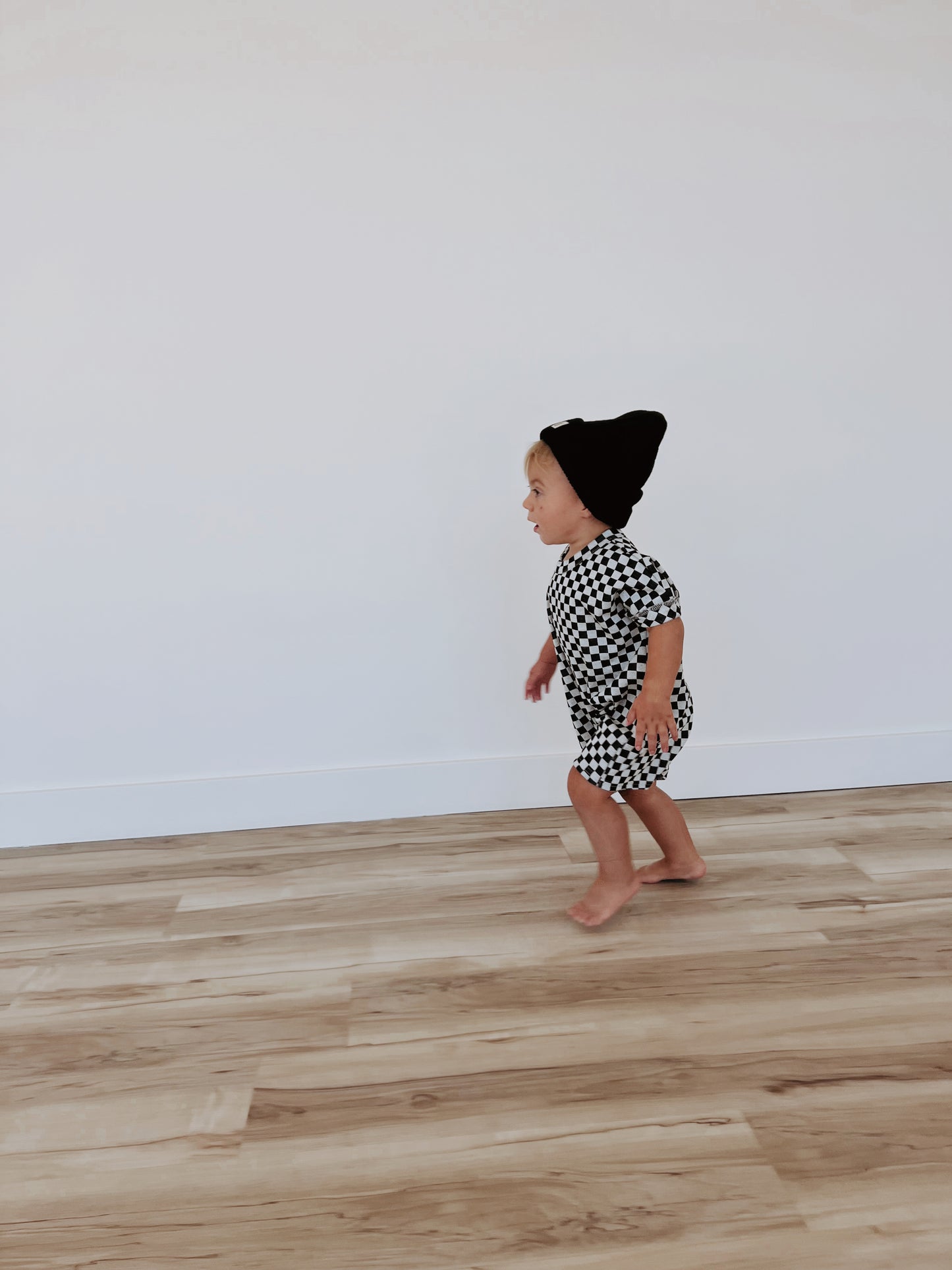 A toddler sporting a Short Sleeve Ribbed Romper in Black & Grey Checkerboard from forever french baby walks barefoot on a light wood floor against a plain white wall. The child, exuding a gender-neutral charm, is captured in profile view with a cheerful expression while looking slightly upwards.