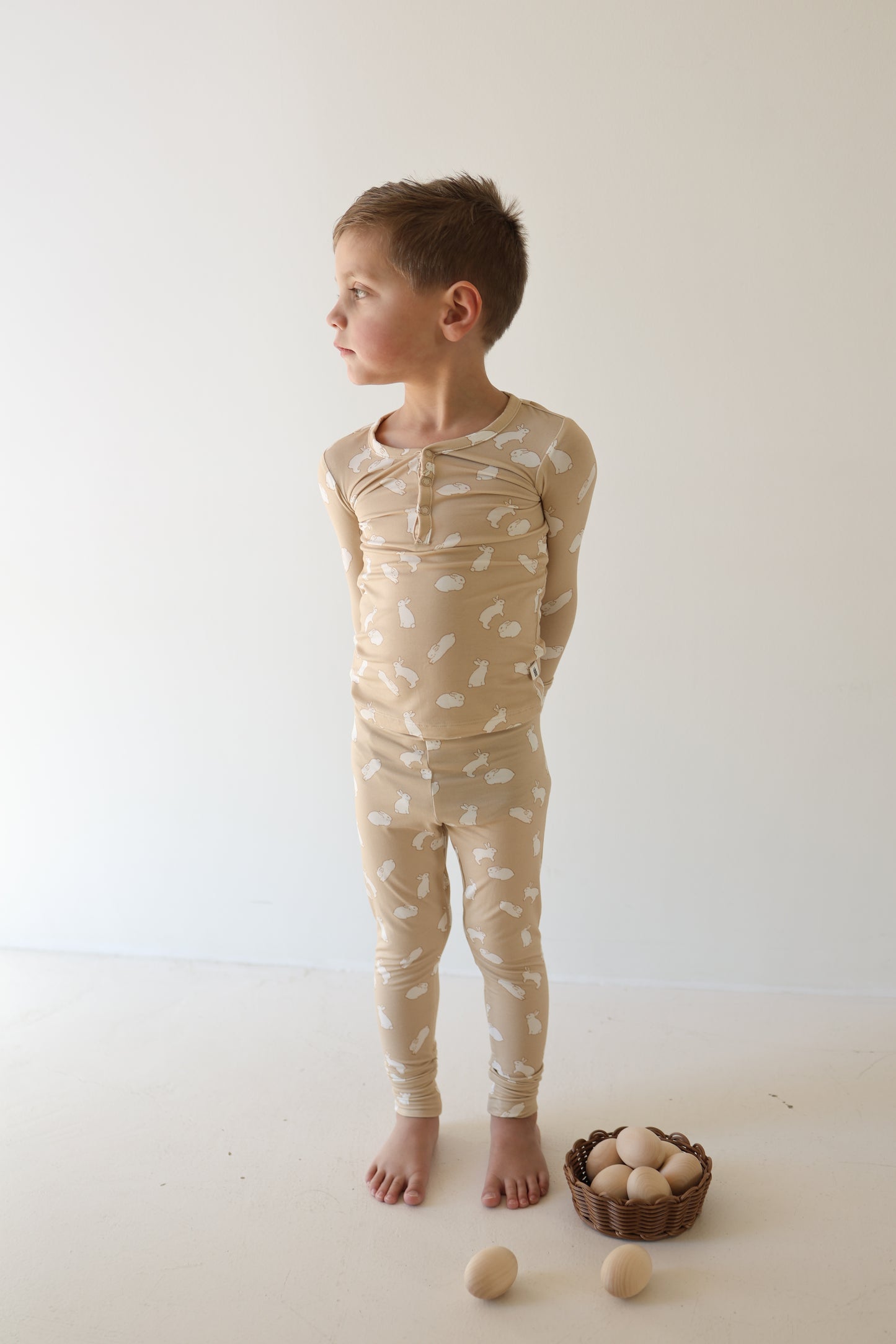 A young child wears forever french baby Bamboo Two Piece Pajamas | Bunnies with white patterns, standing barefoot on a light floor, hands behind back. A small basket of wooden eggs sits nearby against a plain, light background.