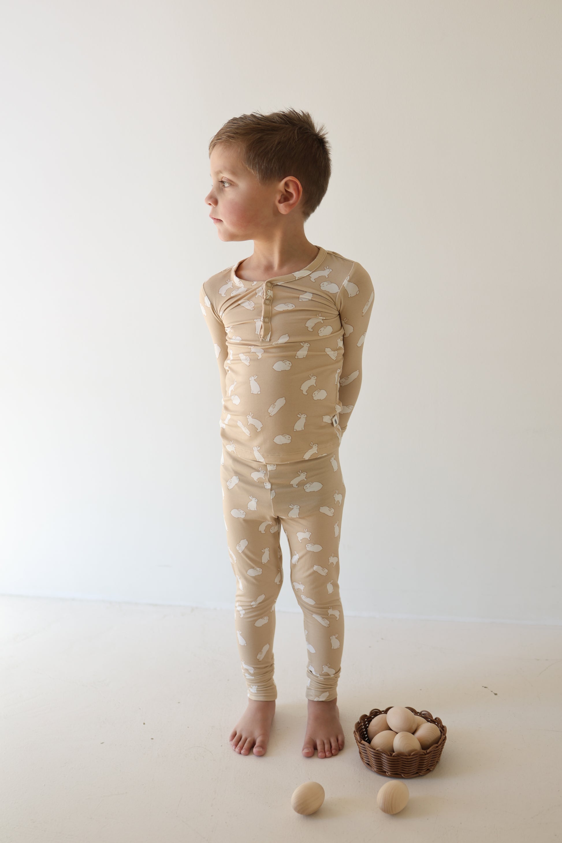 A young child wears forever french baby Bamboo Two Piece Pajamas | Bunnies with white patterns, standing barefoot on a light floor, hands behind back. A small basket of wooden eggs sits nearby against a plain, light background.