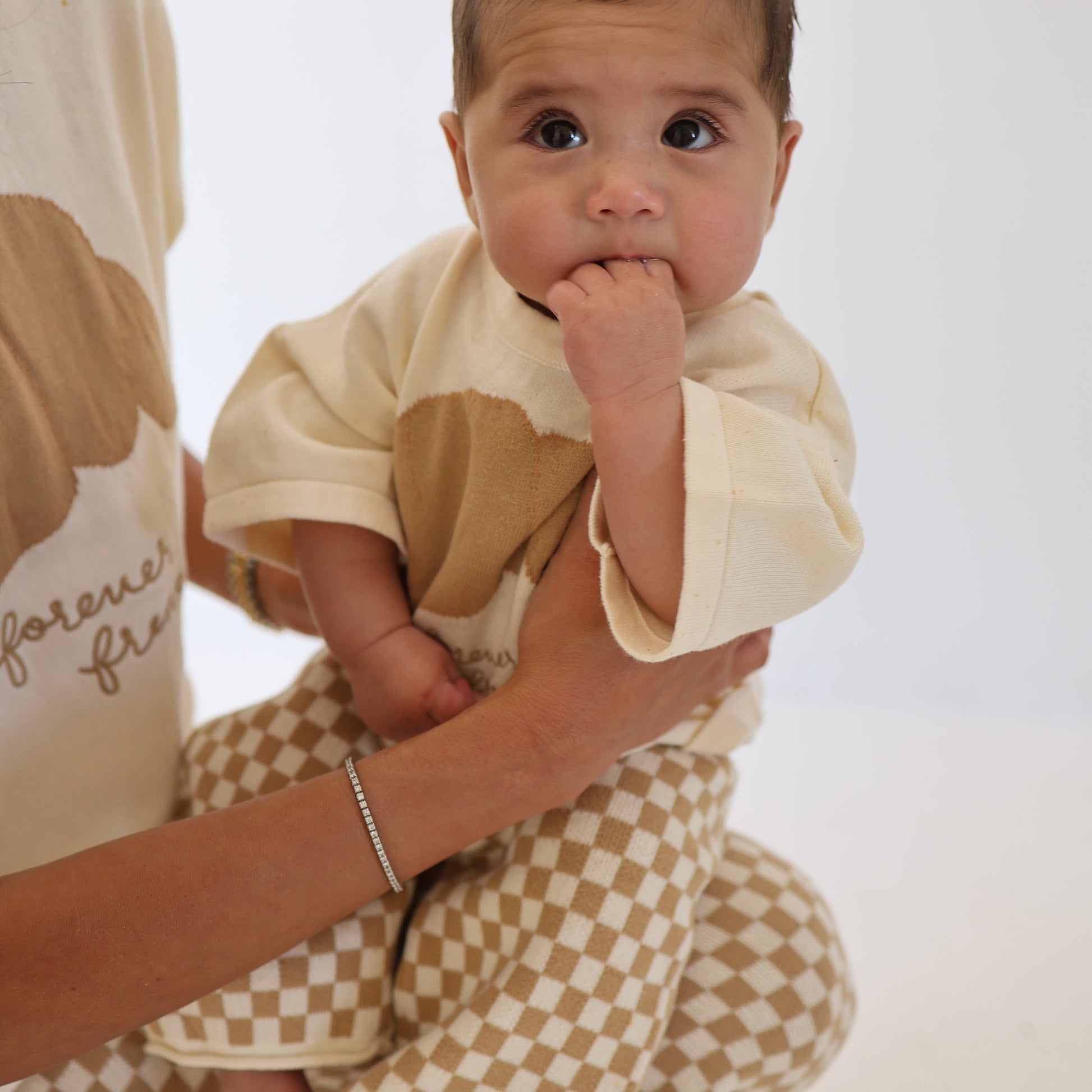 A baby wears the forever french baby Knit Pant Set | Croissants, featuring a cream and tan matching set with checkered pants, while sitting on an adult's lap. The adult has an oversized cloud-patterned top. The background is plain white.