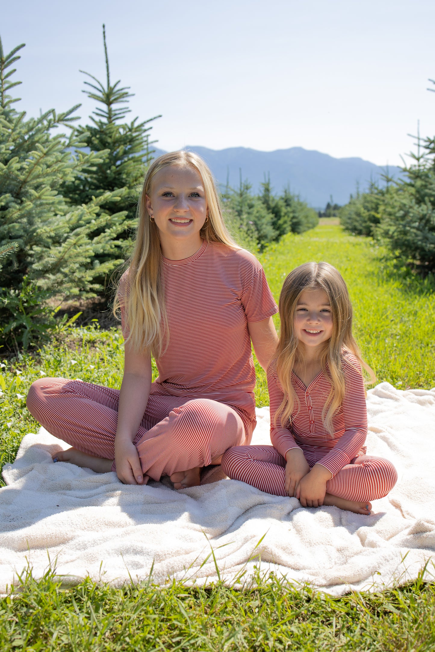 Two girls in matching forever french baby Child Boxy Tee Lounge Sets in Peppermint Stripe Ribbed sit on a blanket outdoors among pine trees. Both are smiling, with mountains and a blue sky visible in the background. Their hypoallergenic kids’ lounge sets keep them comfortable and cozy during their outdoor adventure.