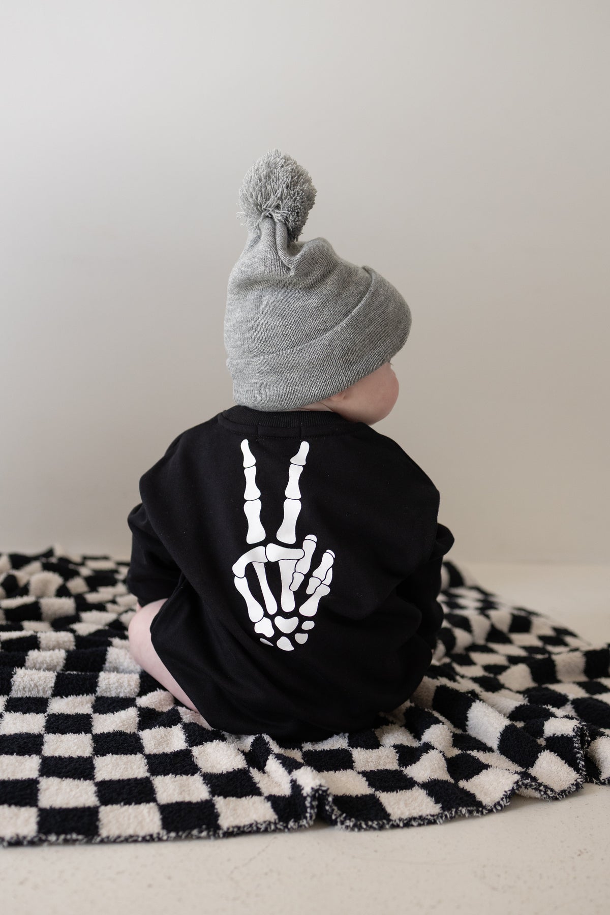 Clad in the forever french baby Sweatshirt Romper | Midnight Skelly—a cozy black outfit featuring a skeleton hand peace sign—a baby wearing a gray beanie sits on a black-and-white checkered blanket, turned away from the camera. Crafted from soft cotton for all-day comfort and cool style, these newborn clothes are perfect for everyday lounging; consult our size chart to ensure the best fit for your little one.