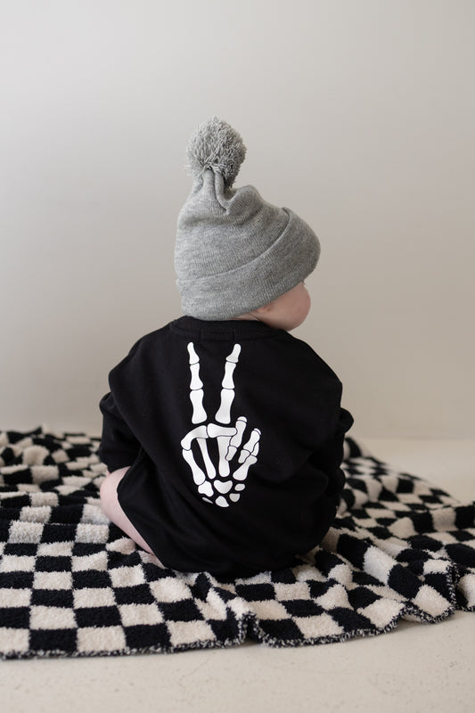 Clad in the forever french baby Sweatshirt Romper | Midnight Skelly—a cozy black outfit featuring a skeleton hand peace sign—a baby wearing a gray beanie sits on a black-and-white checkered blanket, turned away from the camera. Crafted from soft cotton for all-day comfort and cool style, these newborn clothes are perfect for everyday lounging; consult our size chart to ensure the best fit for your little one.