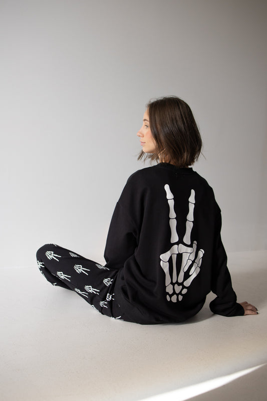 A woman with chin-length brown hair sits on the floor, wearing the forever french baby Women’s Sweat Set | Midnight Skelly, which features bold white skeleton hand designs on the back of the black sweatshirt and on the coordinating pants. She poses sideways against a plain, light background—perfect for embracing spooky season style.
