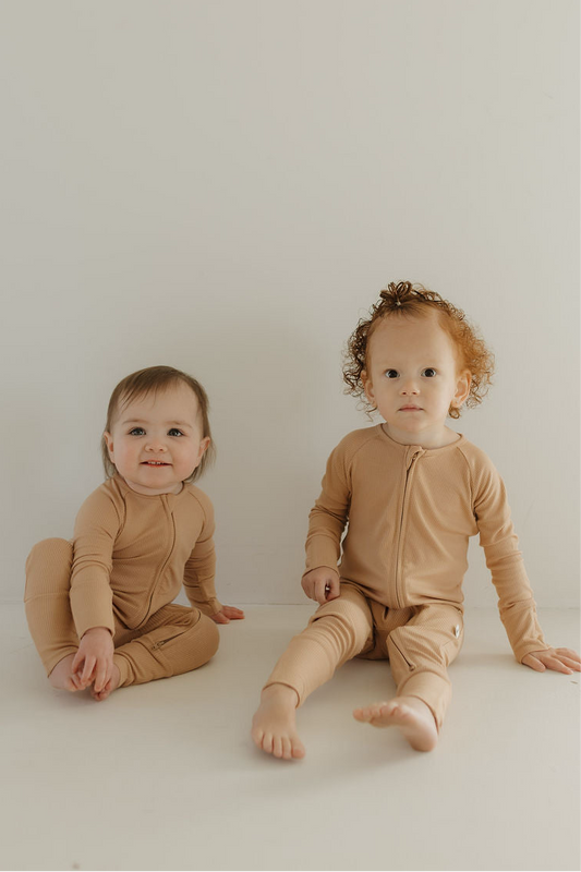 Two toddlers sit by a white wall in matching forever french baby Bamboo Zip Pajamas | Tawny. One smiles with crossed legs, the other sits with legs extended—soft bamboo comfort and style, perfect for all seasons.