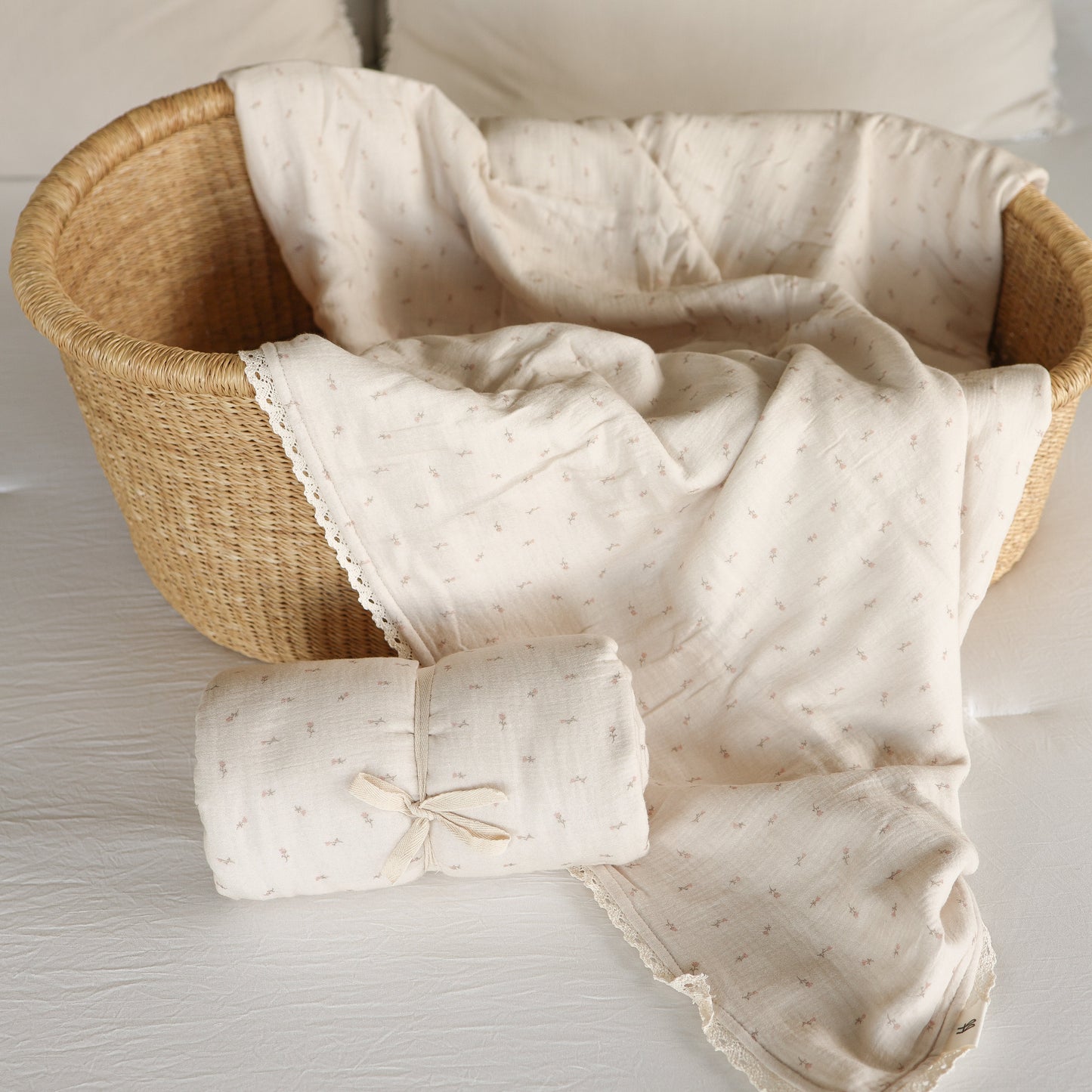 The Muslin Quilt | Lucy by forever french baby features a soft, cream-colored 100% cotton muslin quilt with a subtle pattern in a wicker basket, plus a matching rolled blanket—perfect nursery bedding for your little one.