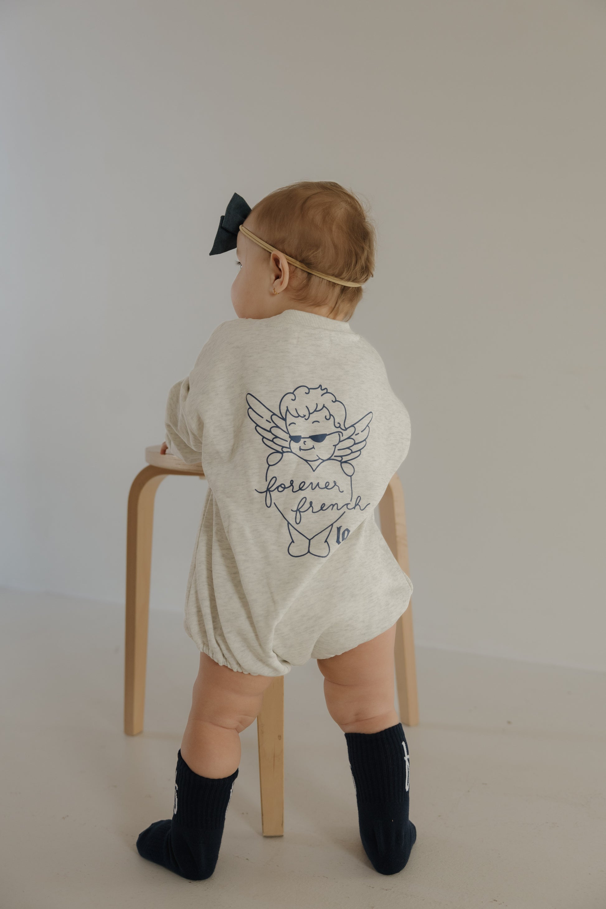 A baby in a cream Sweatshirt Romper by forever french baby (ff Los Angeles), navy knee-high socks, and a large black bow headband stands facing a wooden stool in a minimal room.