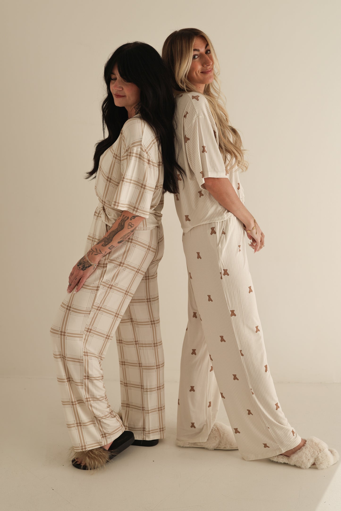 Two women stand back to back, smiling in Women's Boxy Tee Lounge Sets by forever french baby. One wears plaid with dark slippers, the other a small print with light slippers. Their soft bamboo sets are hypo-allergenic, shown on a plain background.