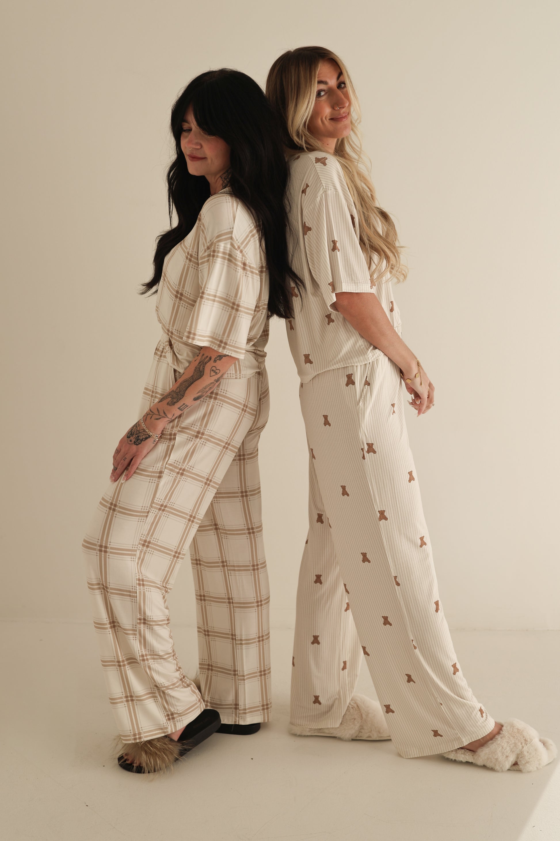 Two women stand back to back, smiling in Women's Boxy Tee Lounge Sets by forever french baby. One wears plaid with dark slippers, the other a small print with light slippers. Their soft bamboo sets are hypo-allergenic, shown on a plain background.