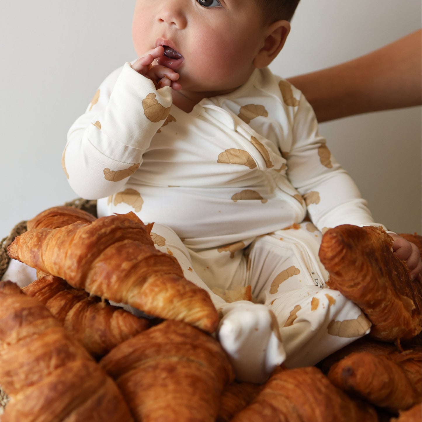 A baby in forever french baby's Bamboo Zip Pajamas | Croissants sits in a basket surrounded by croissants, touching their mouth, with an adult hand supporting them.