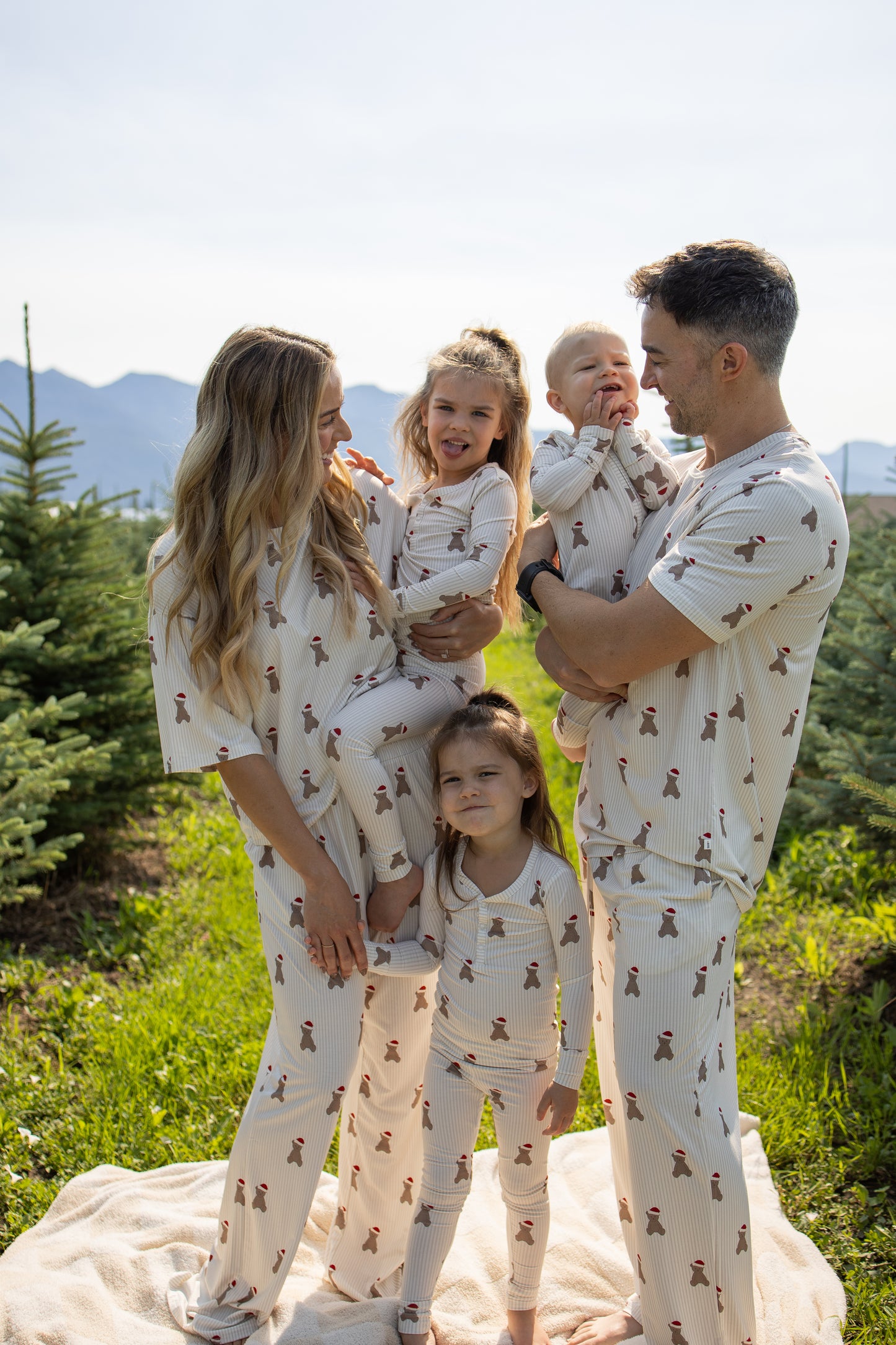 A family of four, all wearing forever french baby's Santa Teddy Men's Short Sleeve Pajamas, stands outdoors on a blanket among pine trees with mountains in the background. The parents each hold a child, and everyone—dressed in matching hypoallergenic pajamas—is smiling and looking at each other.