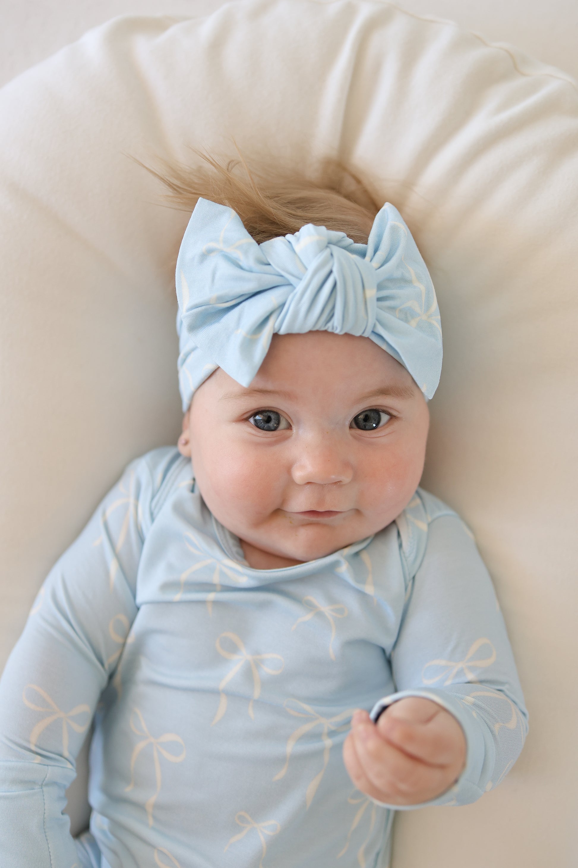 A baby in the forever french baby Long Sleeve Bamboo Snapsuit | Blue Bows and a light blue headband rests on a soft, white cushion, gazing upward with a gentle expression.