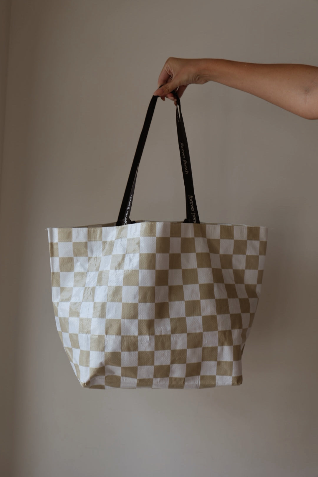 A hand holds up the ff Warehouse Sale Tote by forever french baby, featuring a beige and white checkered pattern with black handles against a plain, light-colored background.