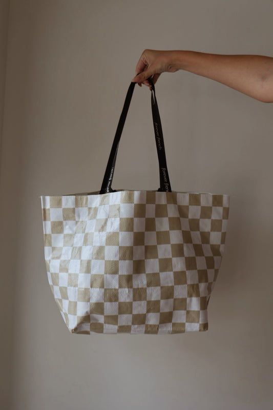 A hand holds up the ff Warehouse Sale Tote by forever french baby, featuring a beige and white checkered pattern with black handles against a plain, light-colored background.