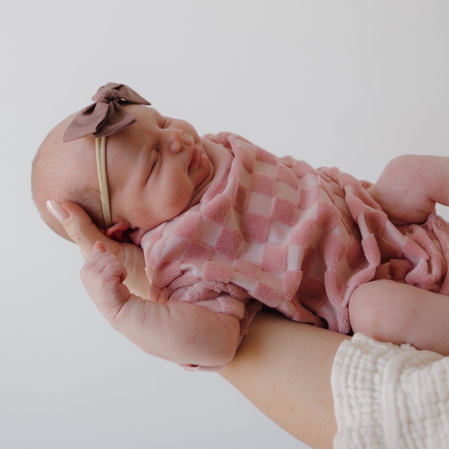 A newborn wearing the forever french baby Terry T-Shirt Romper | Sea Pink is gently cradled in an adult's arms, wrapped in a pink checkered blanket and a mauve bow headband, set against a plain white background.