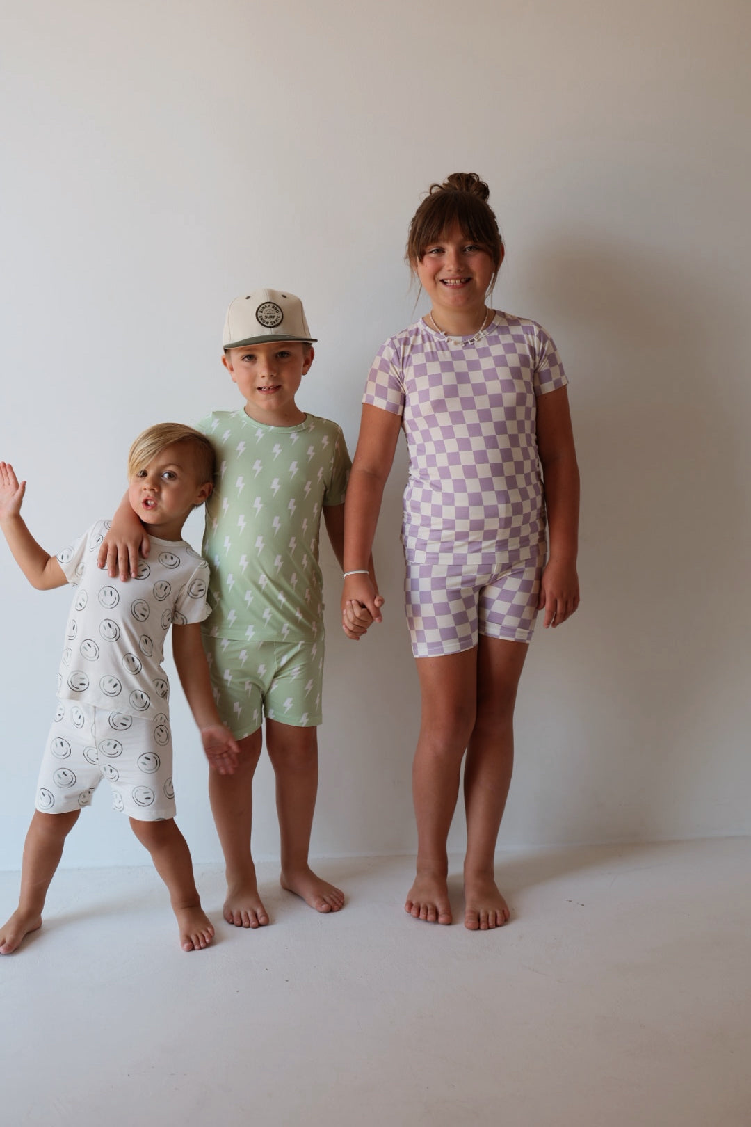Three children stand barefoot against a plain background, holding hands. One wears the forever french baby Bamboo Short Set in Grape Soda—soft, breathable sleepwear that’s perfect for comfy nights.