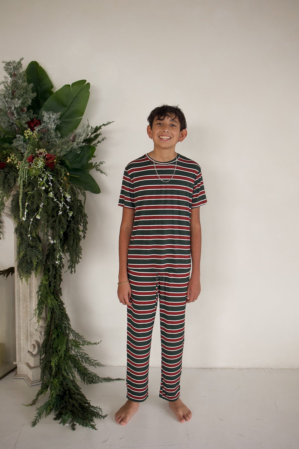 A smiling child stands wearing a pair of Youth Straight Leg Bamboo Pajamas | Holiday Stripe, featuring horizontal red, dark green, and white stripes.