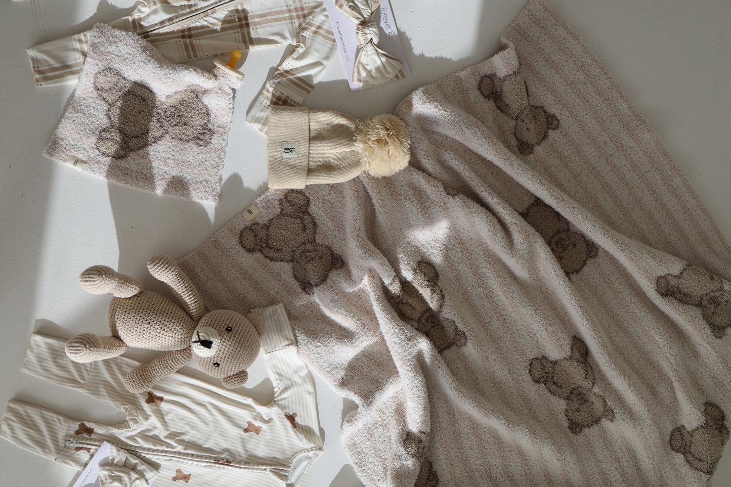 Forever french baby items, including a Plush Blanket | Teddy with teddy bear patterns, a knit teddy bear, a pom-pom hat, and brown motif clothing, all displayed on a white surface in soft daylight.
