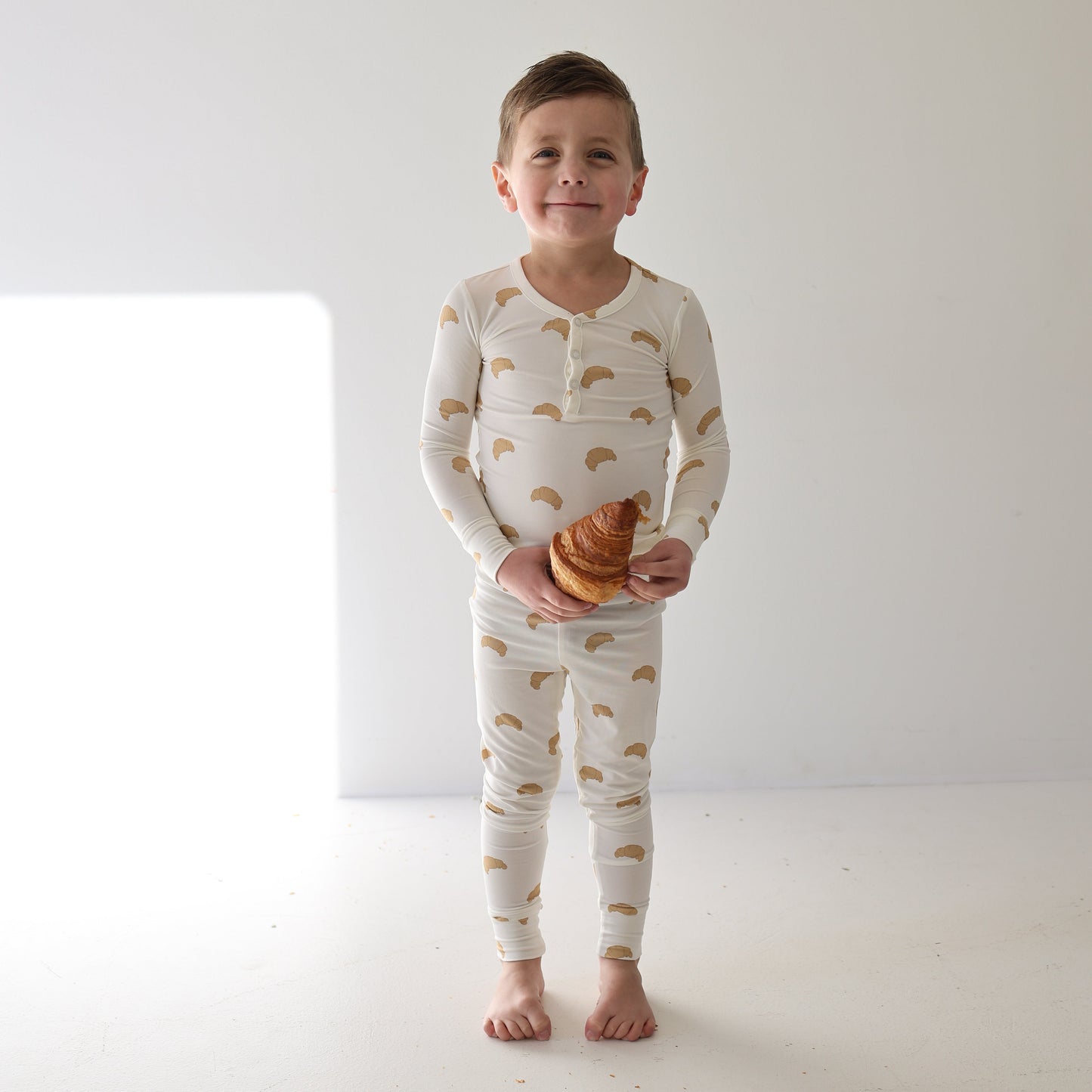 A young boy stands barefoot on a white floor, smiling and holding a croissant while wearing forever french baby Bamboo Two Piece Pajamas | Croissants, with soft light highlighting his breathable sleepwear.
