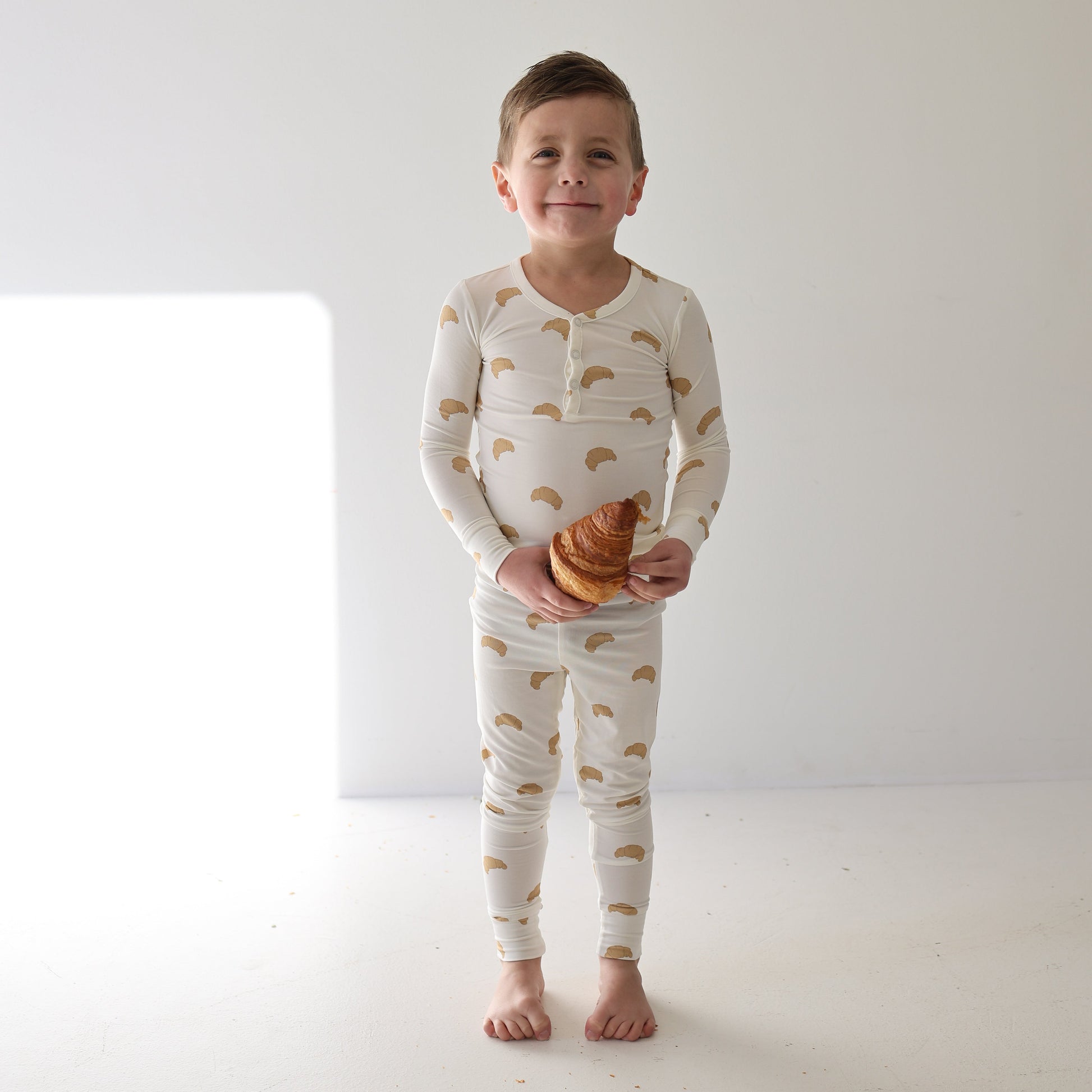 A young boy stands barefoot on a white floor, smiling and holding a croissant while wearing forever french baby Bamboo Two Piece Pajamas | Croissants, with soft light highlighting his breathable sleepwear.