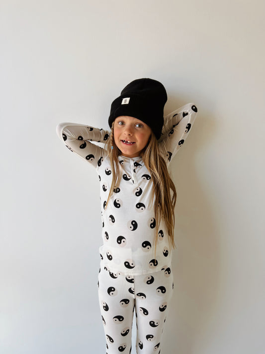 A child wearing forever French baby's Bamboo Two Piece Pajamas in Black & White Yin Yang and a black beanie stands with arms crossed behind their head against a plain background. The child, with long hair, appears to be smiling slightly, showcasing the comfort of the hypo-allergenic clothing.